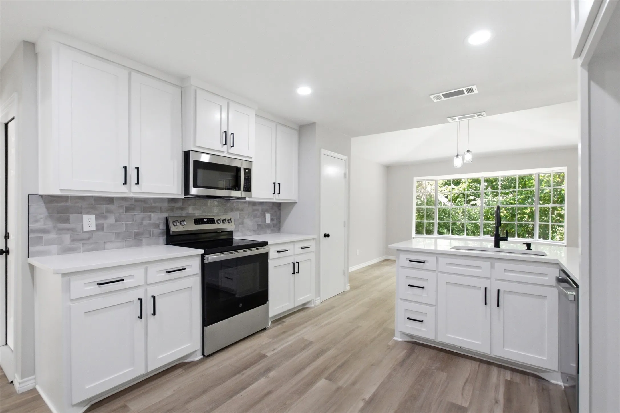 Modern kitchen featuring white cabinetry, stainless steel appliances, light gray wood-finish flooring, a contemporary island with a sink, and a large window overlooking lush greenery