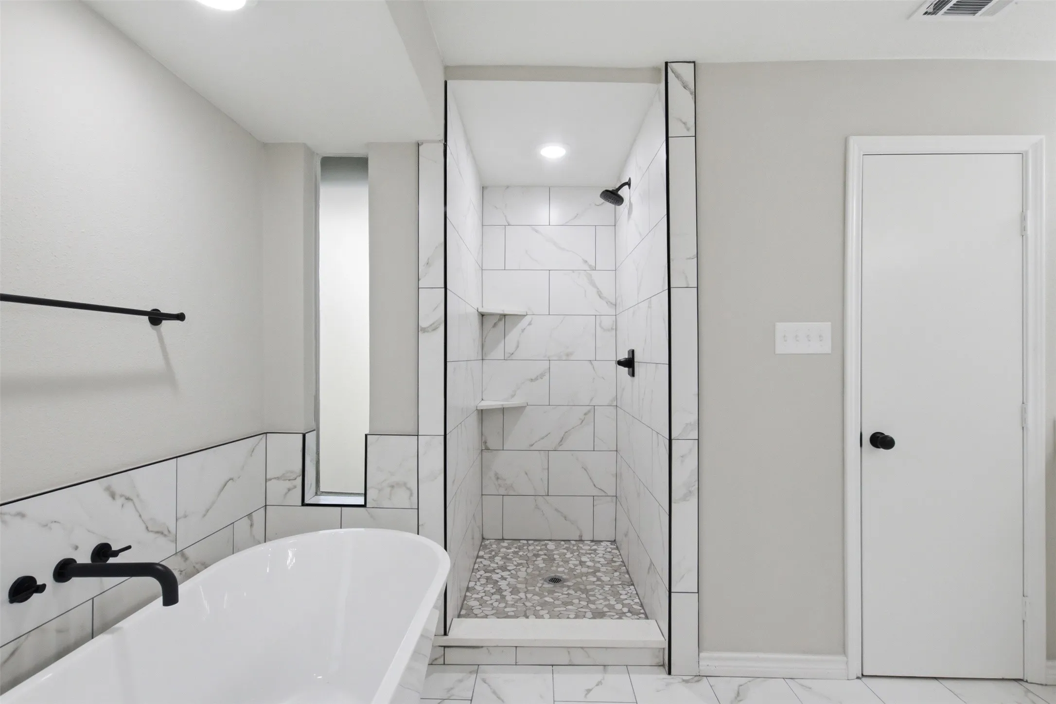 Contemporary bathroom featuring a freestanding soaking tub with wall-mounted matte black fixtures, a walk-in shower with white marbled tile walls, a pebble-finish shower floor, and recessed shelving