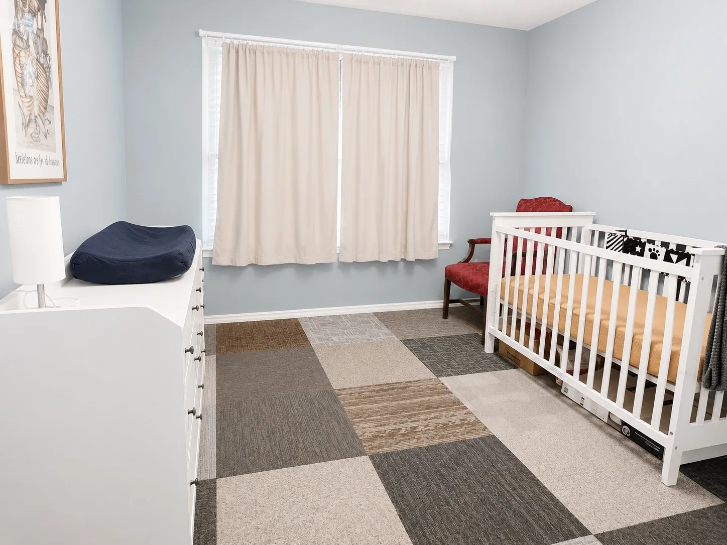 Room featuring light blue walls, a window with blinds and drapes, and multi-tone square carpet tiles