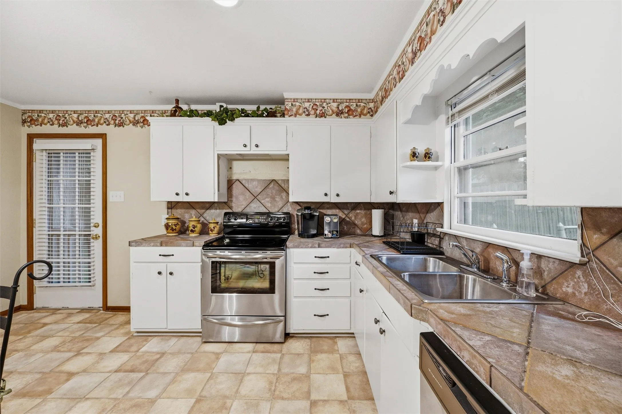 Kitchen features white raised-panel cabinetry with tile countertops and a diagonal tile backsplash. Stainless steel electric range and dishwasher are included. Double sink sits beneath a window overlooking the backyard. Tile flooring extends throughout.