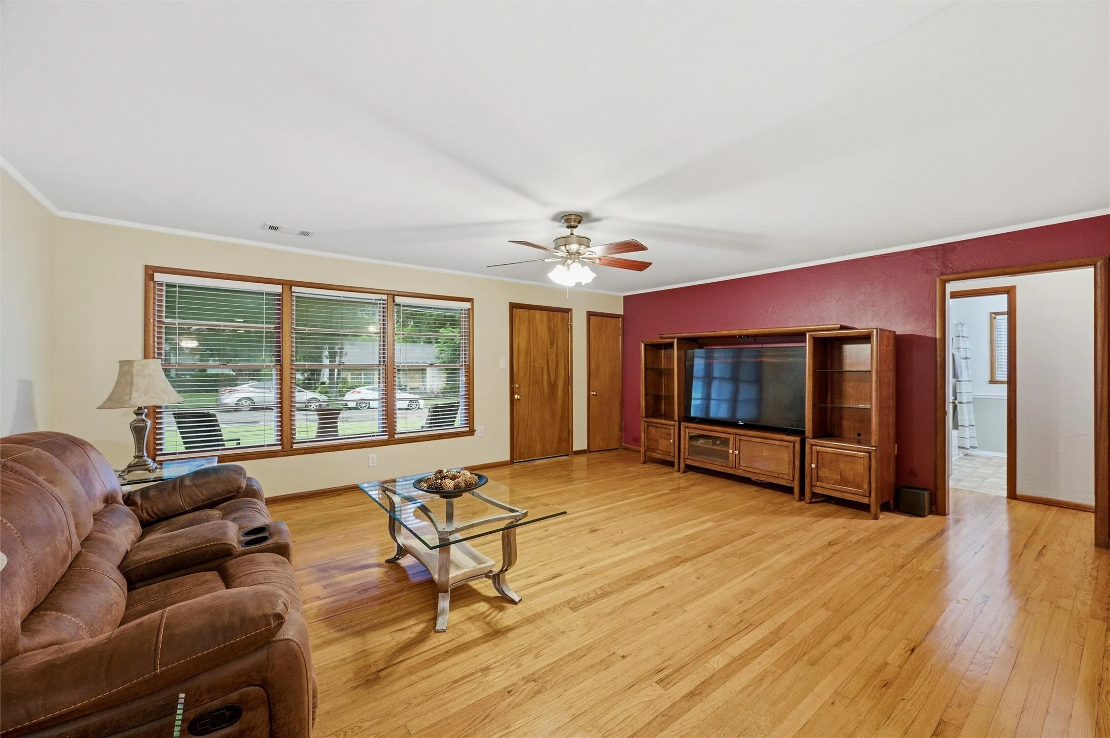 Living Room features crown molding and a ceiling fan with light fixture. Large picture window fills the room with natural light. Deep red accent wall provides contrast against cream-painted walls. Hardwood flooring extends throughout.