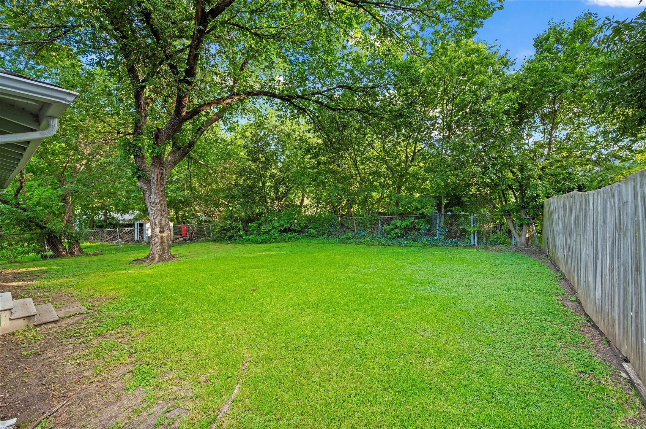 Backyard features an open lawn surrounded by mature trees and landscaping. Chain-link fencing encloses the rear yard with wood privacy fencing along the side.