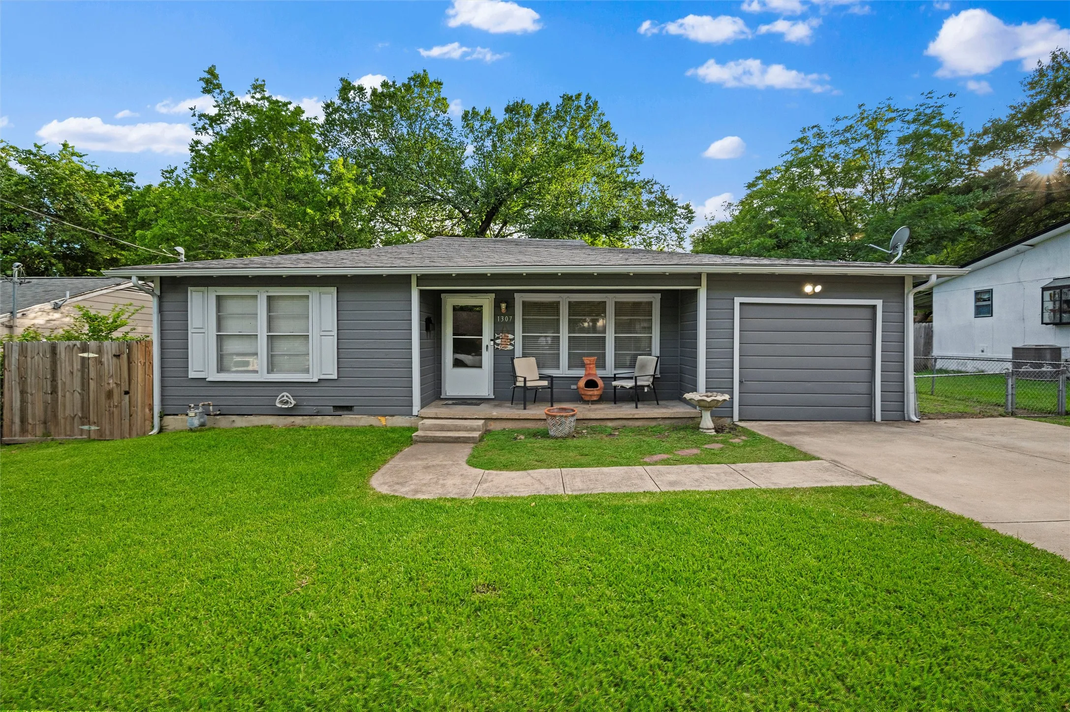 Front Exterior features gray lap siding with white trim and shutters. Covered front porch connects to the attached garage. Concrete driveway and walkway provide front access. Mature trees enhance the front yard.