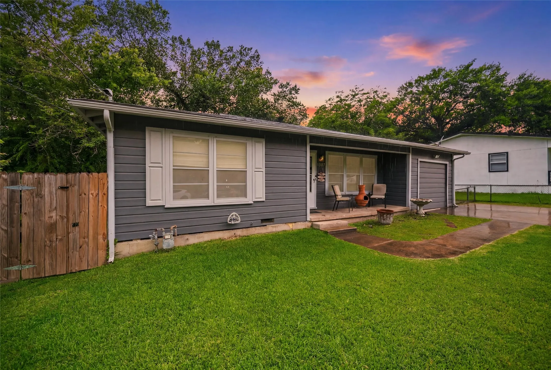 Front Exterior features gray lap siding accented by white trim and shutters. Covered front porch spans the entry alongside a single-car attached garage. Concrete walkway connects to the driveway. Mature trees frame the property.