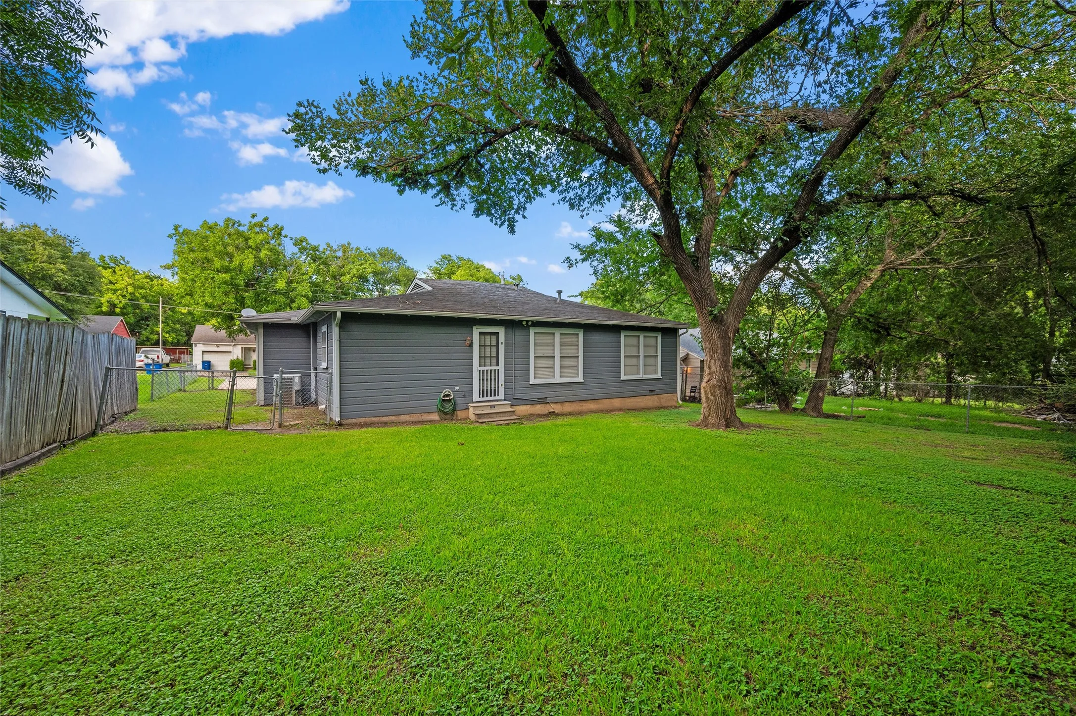 Rear Exterior features gray lap siding with white-trimmed windows and rear entry door. Mature shade tree provides backyard shade. Wood privacy and chain-link fencing enclose the outdoor space.
