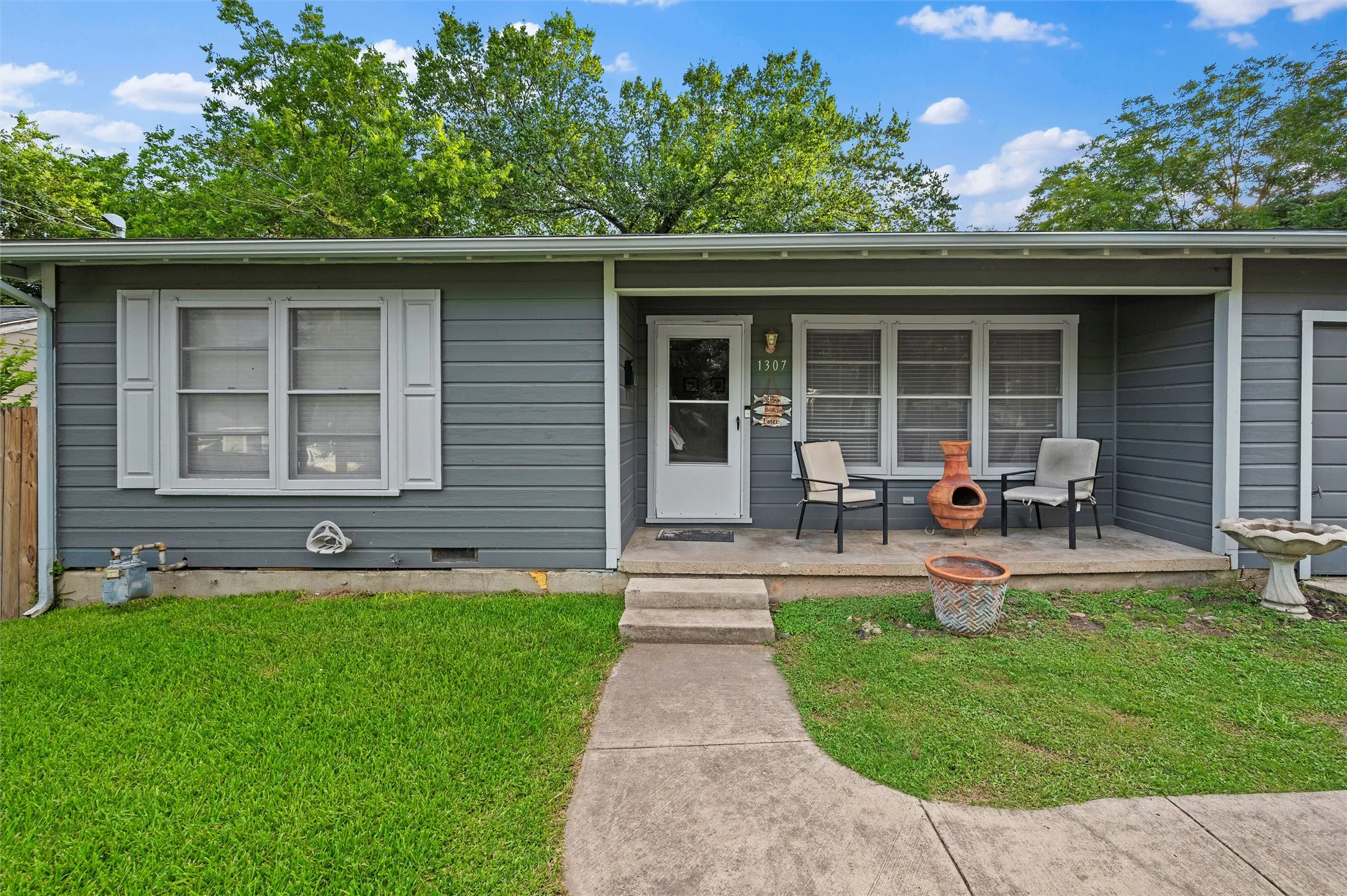 Front Entry features a covered concrete porch leading to the front door. Gray lap siding, white-trimmed windows, and shutters frame the entrance. Address numbers are visible at the entry.