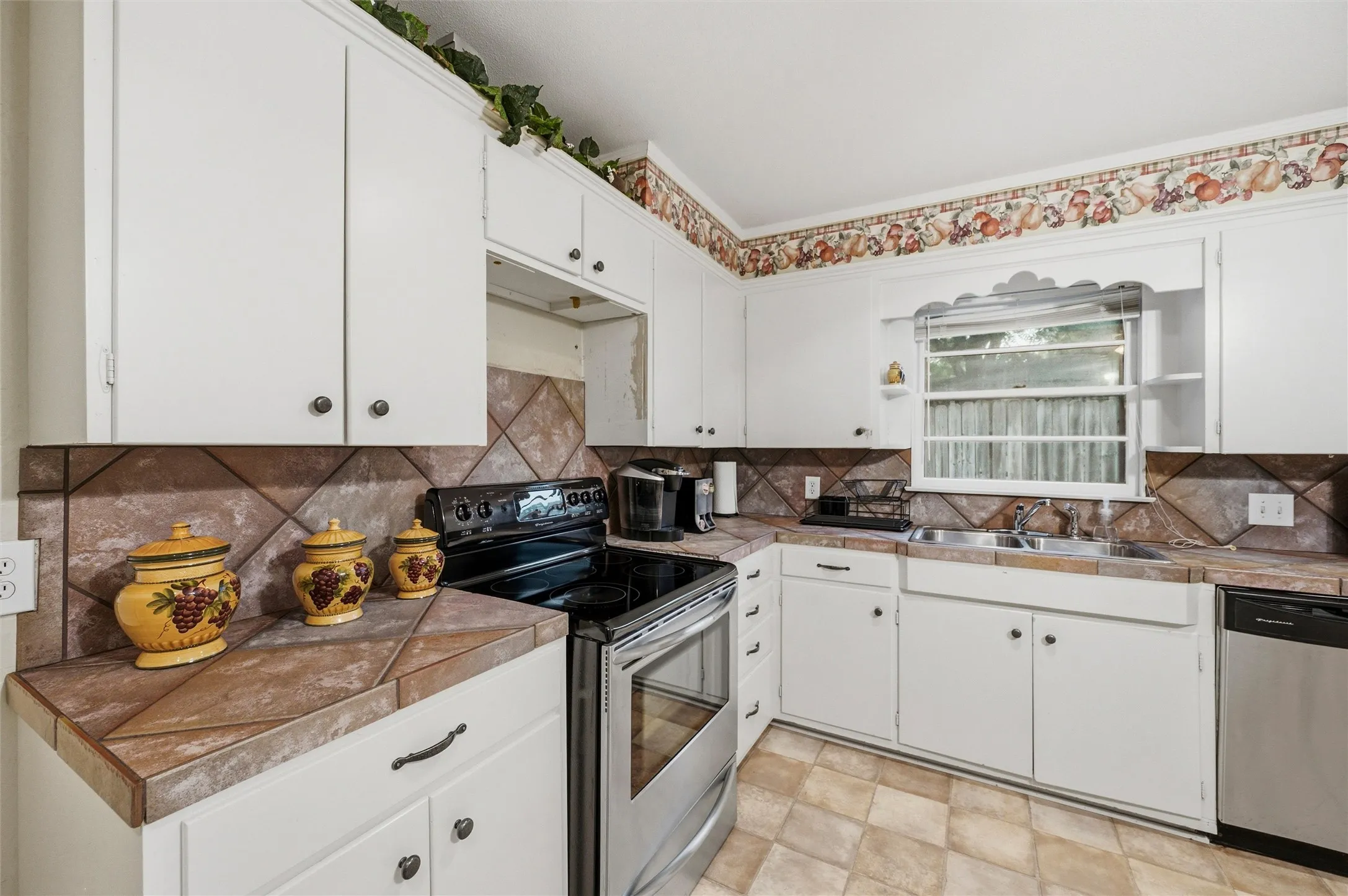 Kitchen features white raised-panel cabinetry, tile countertops, and a diagonal tile backsplash. Stainless steel electric range and dishwasher complement the workspace. Double sink overlooks the backyard. Tile flooring extends throughout.