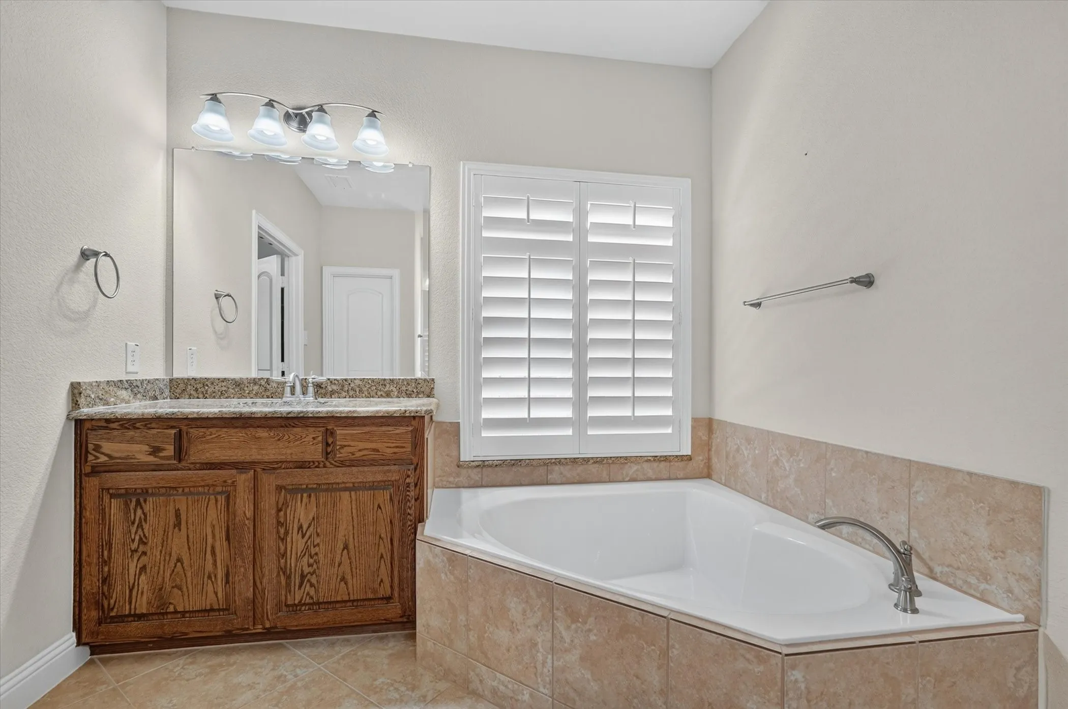 Bathroom featuring a corner soaking tub with tile surround, wood-finish vanity with countertop, and window with plantation shutters