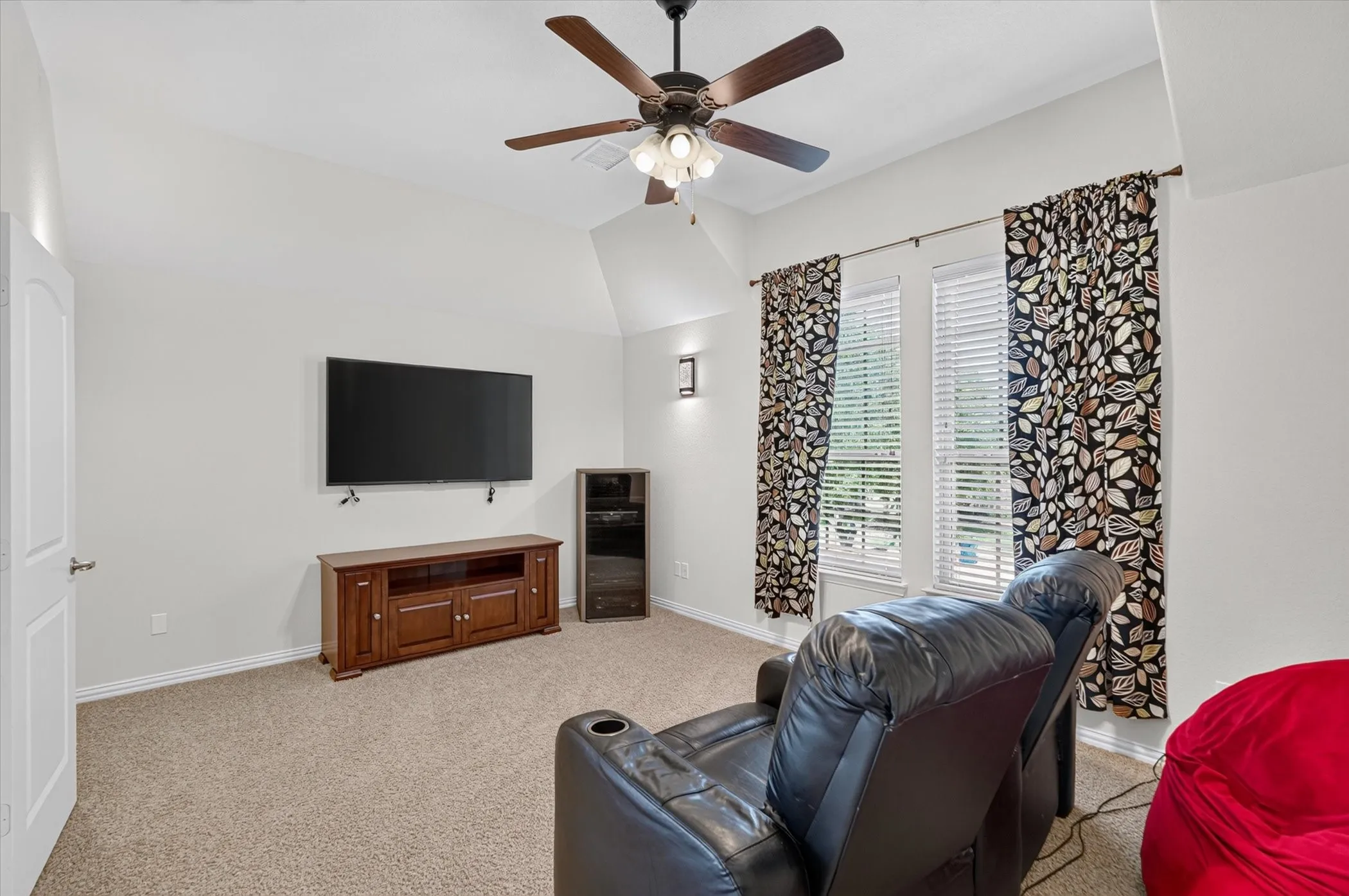 Room with vaulted ceiling, ceiling fan with integrated lighting, wall-mounted television, wood-finish media console, and two windows with blinds and patterned curtains