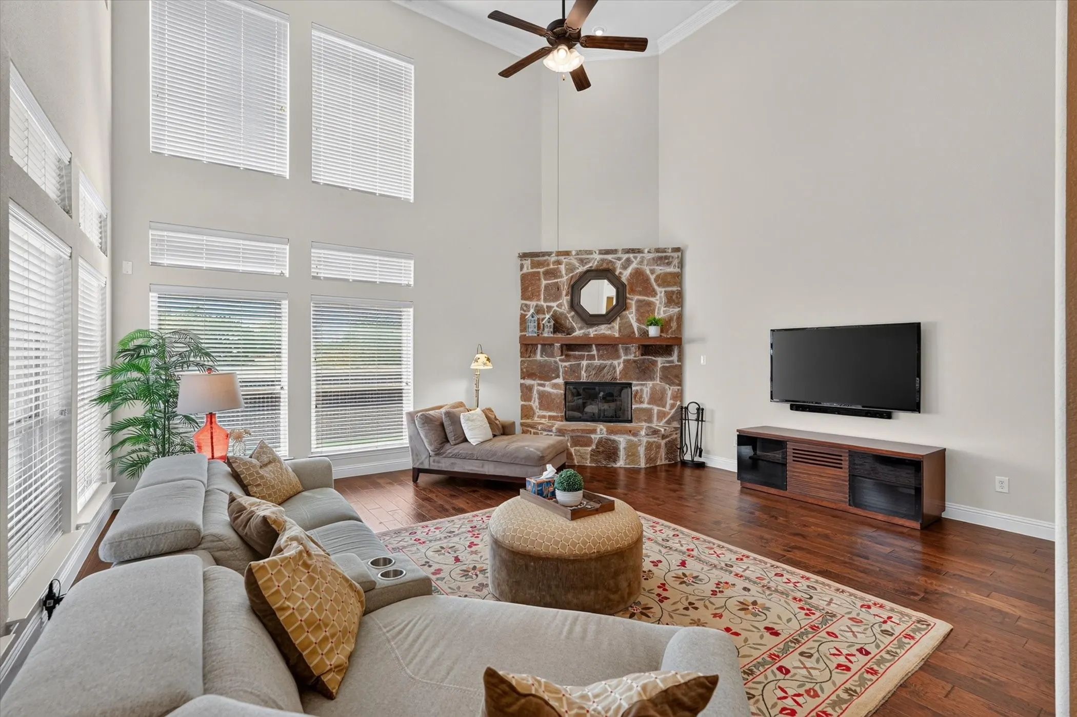 Vaulted ceiling with a ceiling fan, stone fireplace with a wood mantel, ample natural light through tall windows, and rich wood-finish flooring