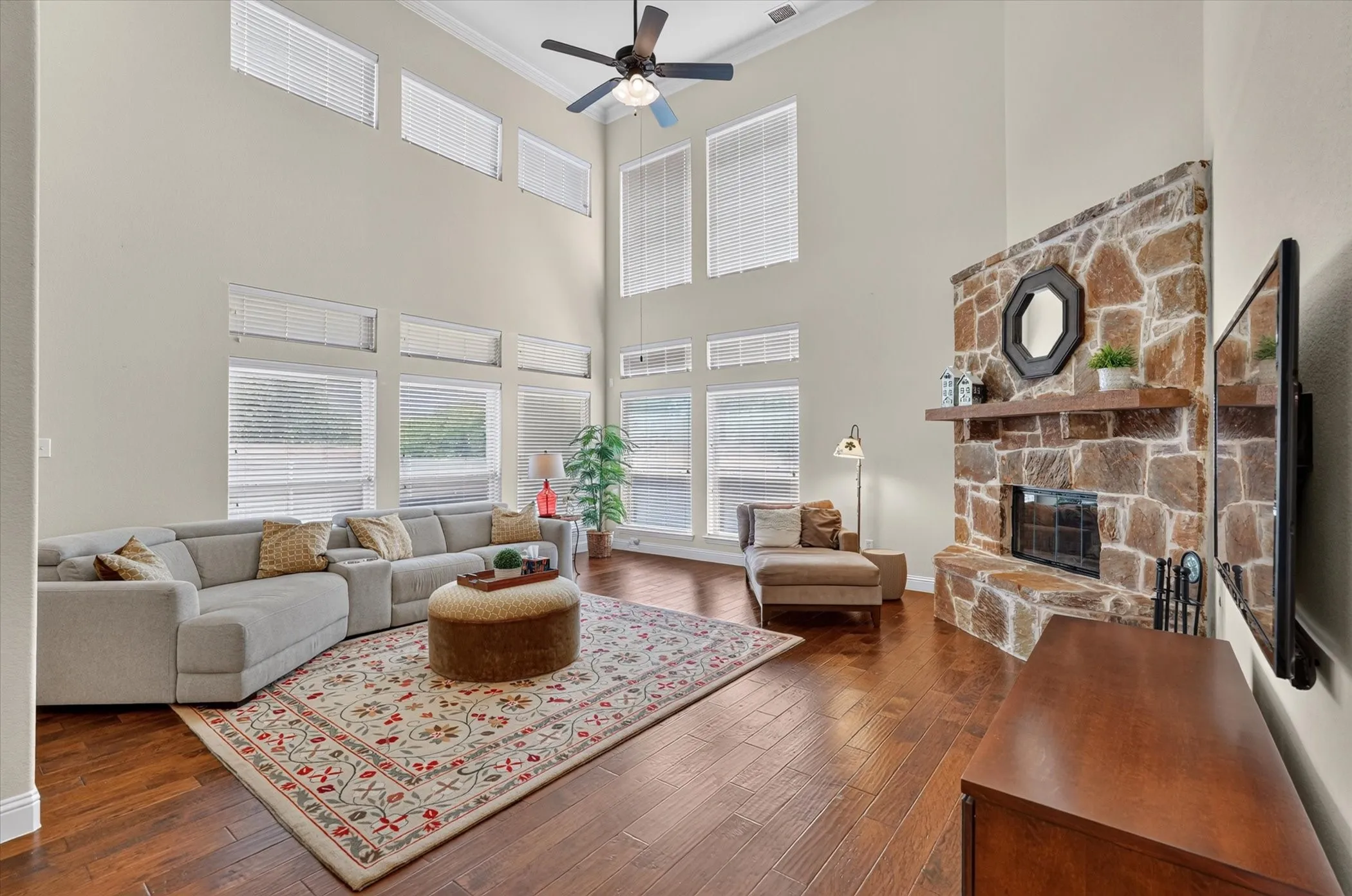 Spacious living area featuring a soaring vaulted ceiling, extensive window array with upper transom windows, a stone-clad fireplace with a solid wood mantel, wood-finish flooring, and a ceiling fan