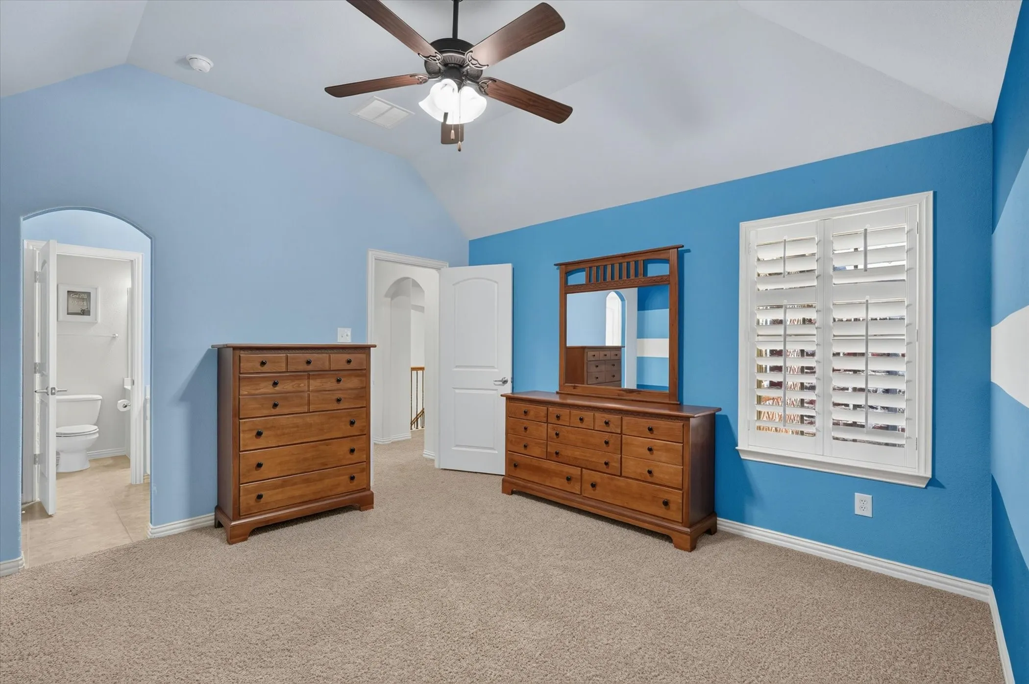 Vaulted ceiling room featuring carpet flooring, a ceiling fan with light, a window with white plantation shutters, and an arched doorway