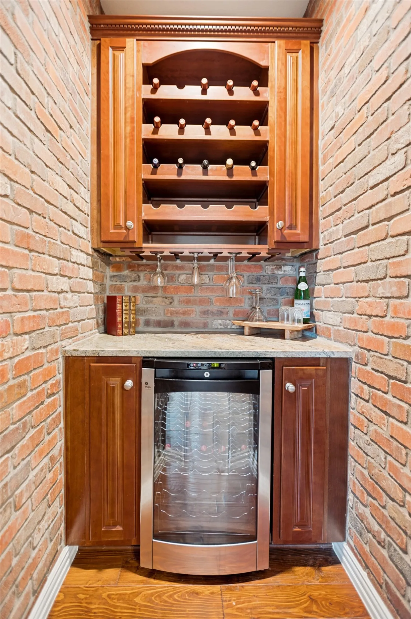 Built-in beverage station featuring exposed brick walls, wood-finish flooring, cherry-toned cabinetry, a granite countertop, and an integrated beverage refrigerator