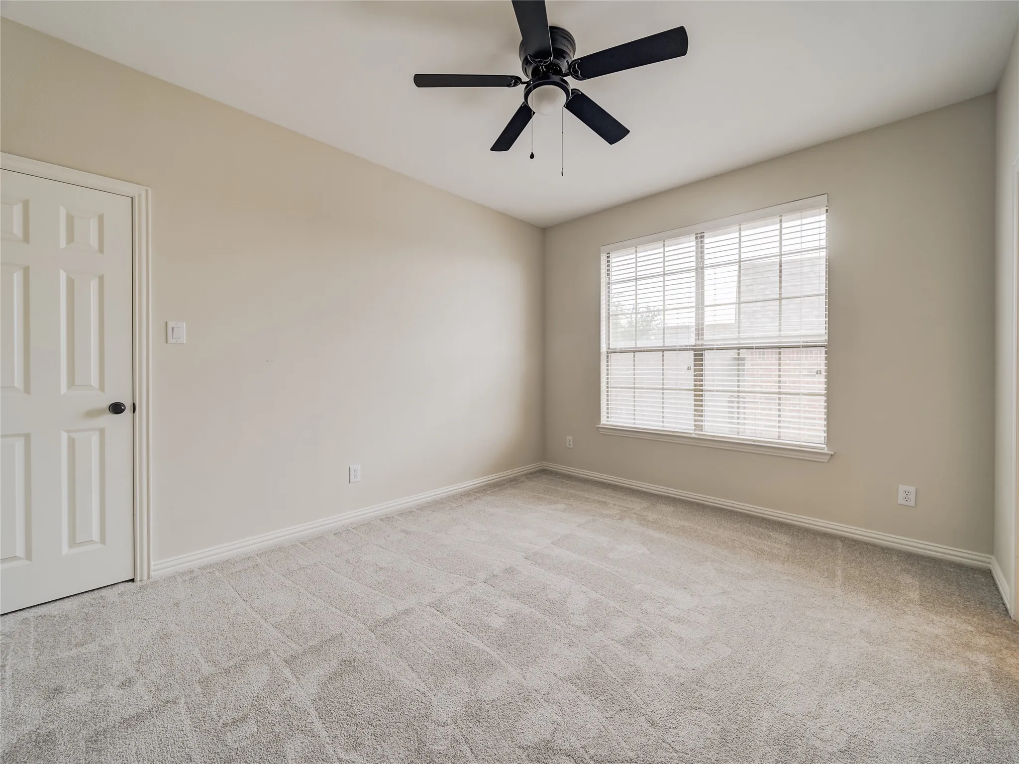 Spacious room featuring neutral wall paint, light-toned carpeting, a multi-pane window with blinds, and a black ceiling fan with an integrated light fixture