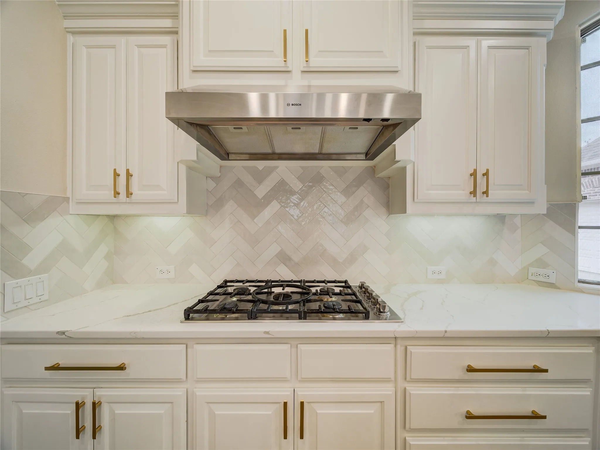 Gourmet kitchen featuring a stainless steel range hood, built-in gas cooktop, white shaker-style cabinetry with gold hardware, herringbone pattern backsplash, and stone countertops