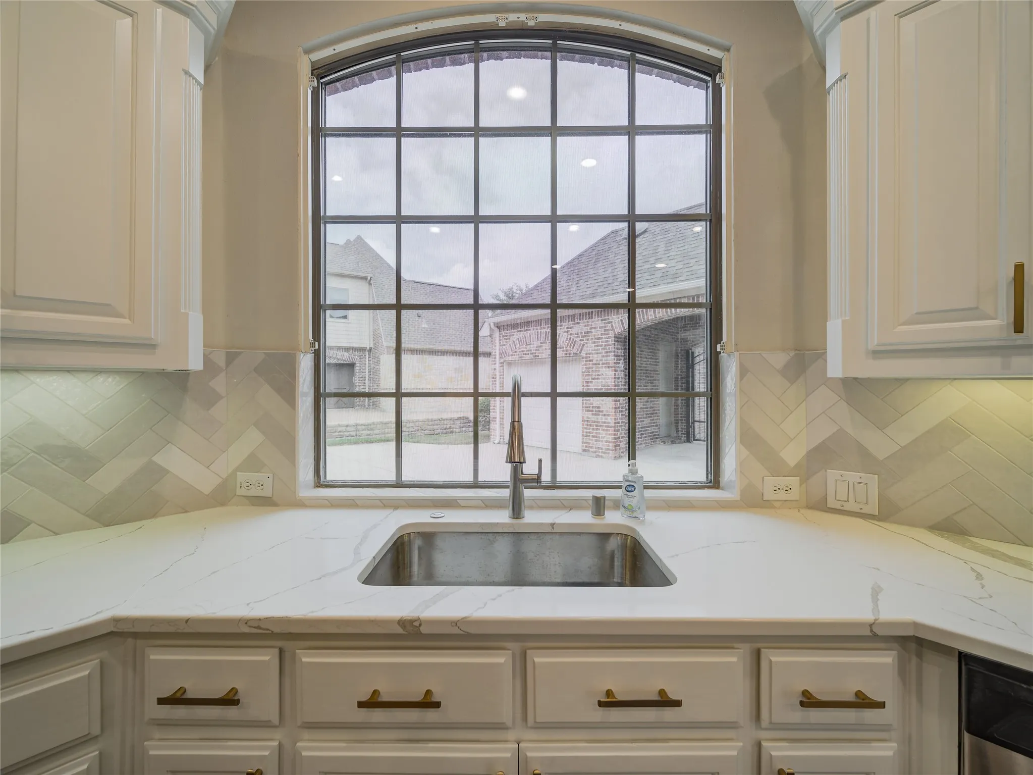 Kitchen sink area featuring an arched window with grid panes, white cabinetry with gold-toned hardware, light countertops, and a herringbone pattern tile backsplash