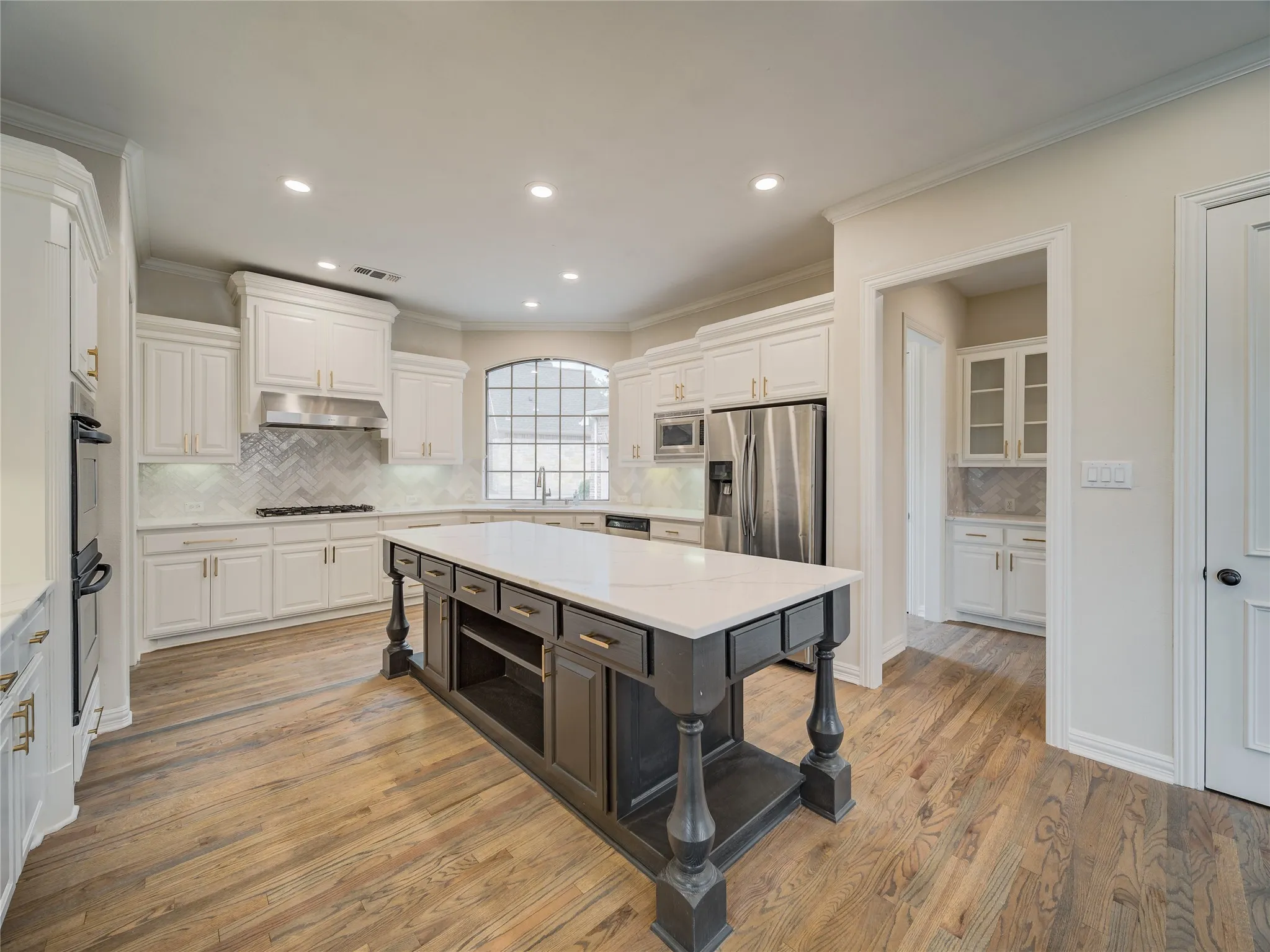 Spacious kitchen featuring a central island with a light-toned countertop, white cabinetry with gold-toned hardware, stainless steel appliances, herringbone-patterned backsplash, and wood-finish flooring