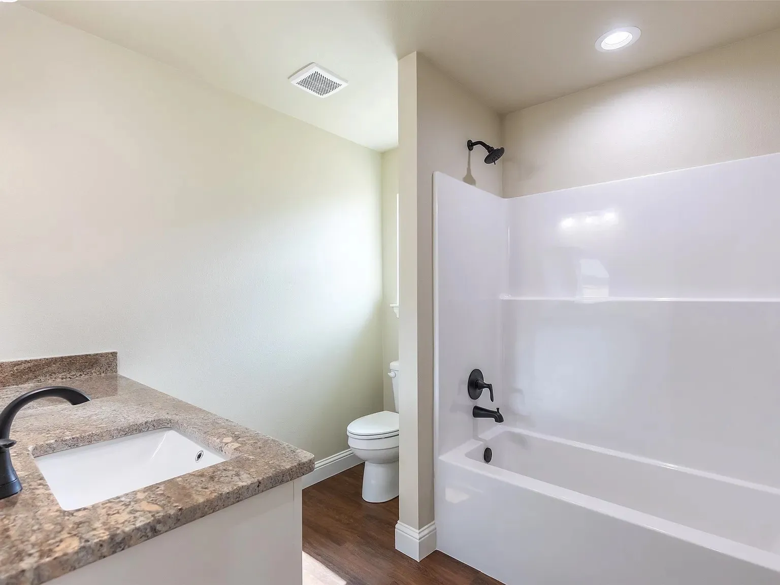 Full bathroom featuring a white tub/shower combination with matte black fixtures, a single vanity with a granite-style countertop, and wood-finish flooring