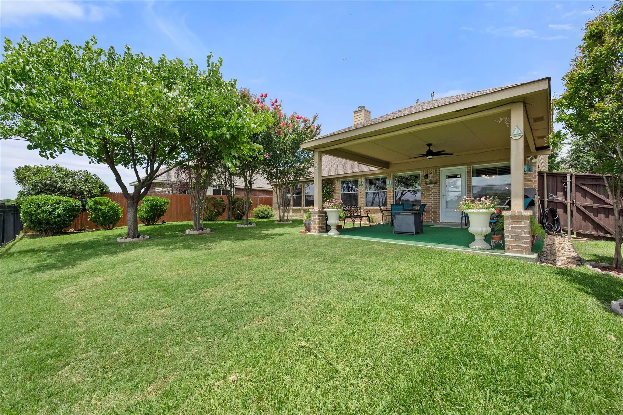 This beautifully landscaped backyard retreat showcases the home's inviting rear elevation with a large covered patio, creating the perfect space for outdoor living and entertaining. The expansive fully fenced backyard features a lush, well-maintained lawn, mature shade trees, and attractive landscaping that provide both beauty and privacy. The oversized covered patio includes a ceiling fan and ample space for outdoor dining and relaxing while enjoying peaceful backyard views. With plenty of room for children, pets, gardening, or future outdoor enhancements, this private backyard offers the ideal combination of comfort, functionality, and curb appeal, making it a standout feature for buyers seeking exceptional Texas outdoor living.