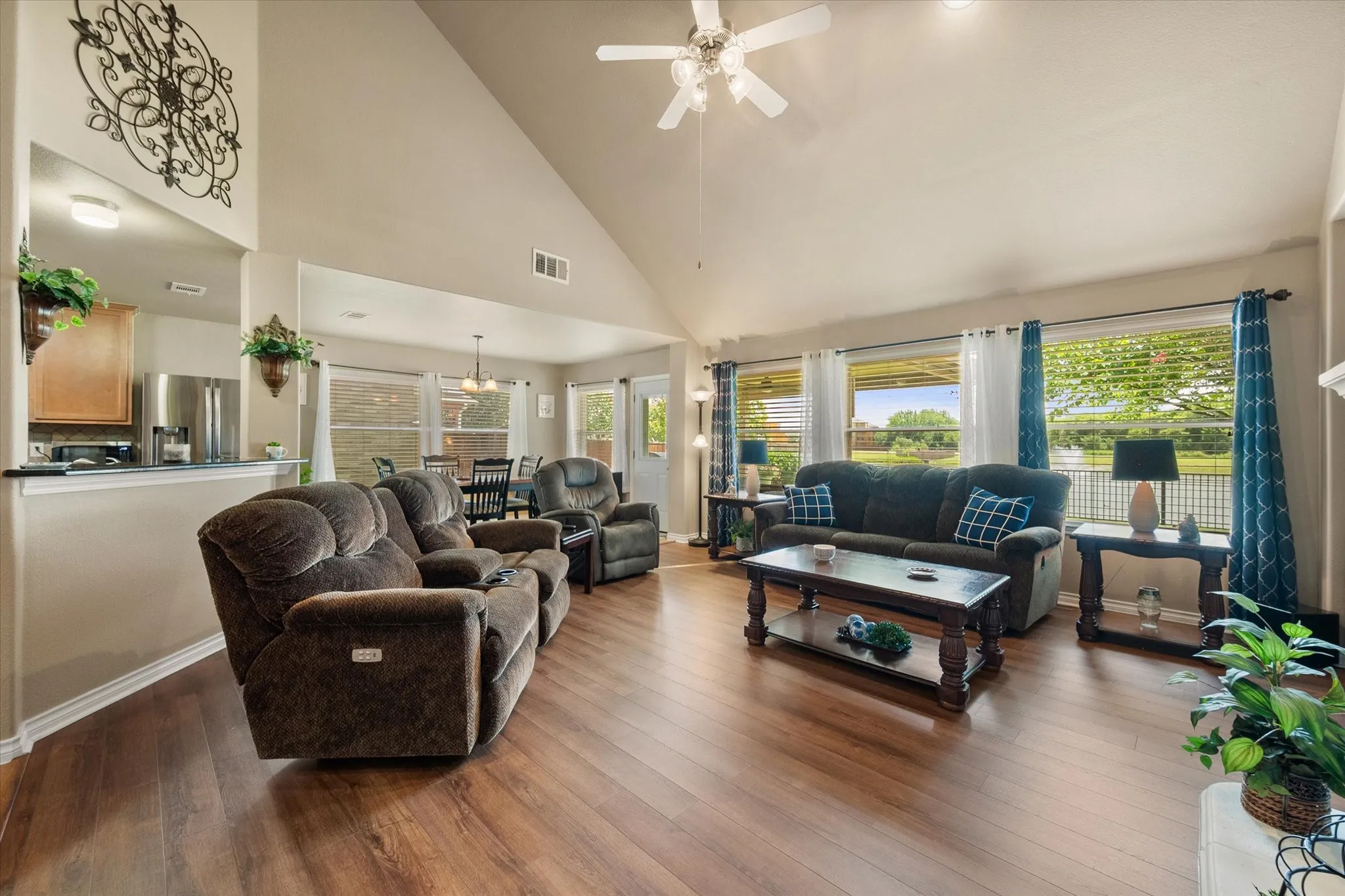 This spacious open-concept living room offers the perfect blend of comfort and style with soaring vaulted ceilings, rich warm-toned luxury vinyl plank flooring, and an abundance of natural light. The expansive layout flows seamlessly into the dining area and kitchen, creating an ideal space for entertaining and everyday living. Neutral designer finishes, warm color palettes, and the airy floor plan enhance the home's inviting atmosphere, while oversized windows frame peaceful backyard views and bring the outdoors in. This beautifully designed gathering space is both functional and welcoming, making it the heart of the home.