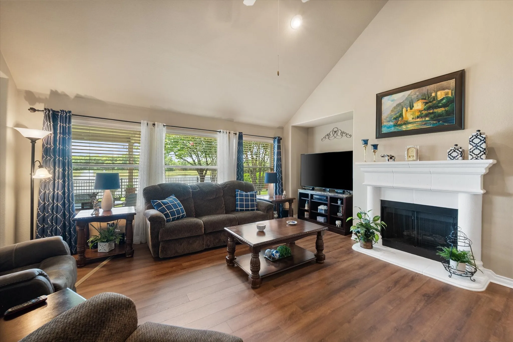 This impressive living room showcases luxury vinyl plank flooring, soaring vaulted ceilings, and a dramatic wall of windows that fills the space with natural light while offering peaceful views of the lush, landscaped backyard. The open, airy design creates an inviting atmosphere perfect for everyday living and entertaining. The expansive windows seamlessly connect the indoor living space with the outdoor scenery, enhancing the home's bright and spacious feel. Neutral finishes and an open-concept layout make this room both stylish and versatile.