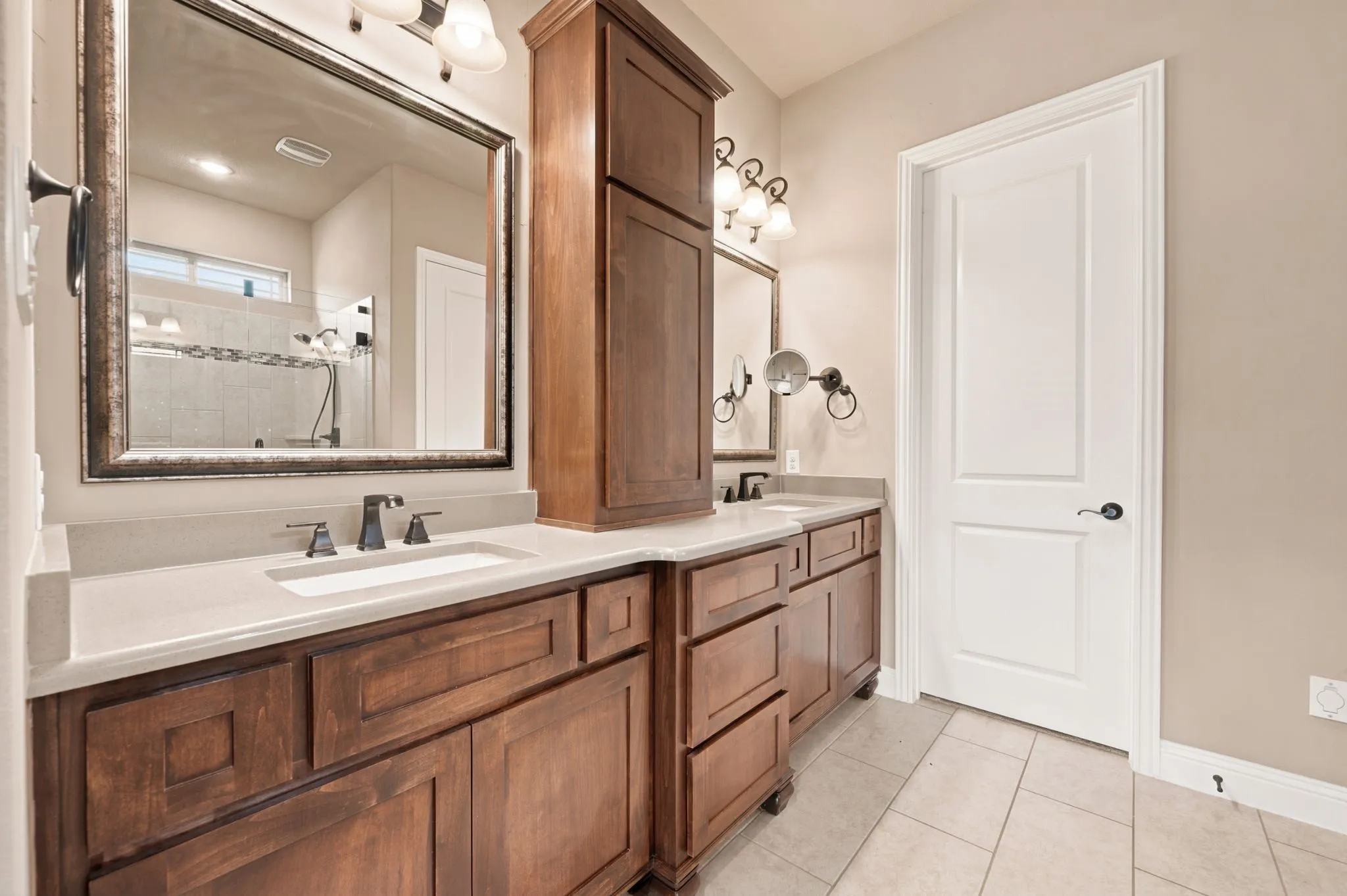 Dual undermount sink vanity includes a quartz counter top, custom built cabinetry, framed mirrors and a medicine cabinet. Bronze fixtures. Floors are tiled. Door leads into a custom designed wardrobe closet.