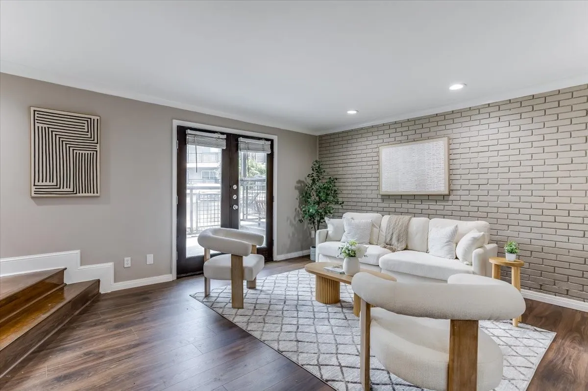 Living area featuring wood-finish flooring, a light-toned brick accent wall, recessed lighting, and double doors opening to an exterior space