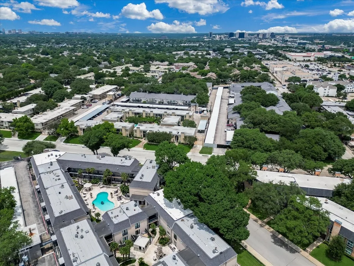 Community aerial featuring multiple residential buildings with pitched roofs, mature trees, a central swimming pool with surrounding patio, and covered parking structures
