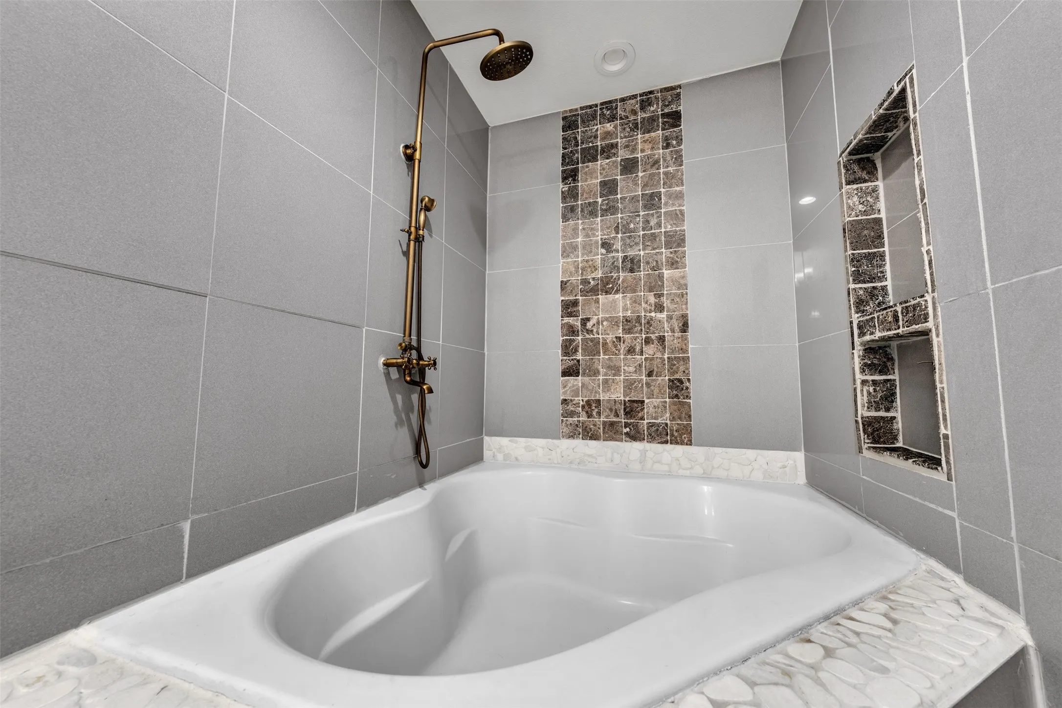 Freestanding soaking tub featuring a bronze-finish shower fixture, light gray wall tiles, a mosaic tile accent strip, and a recessed shower niche with matching mosaic tile