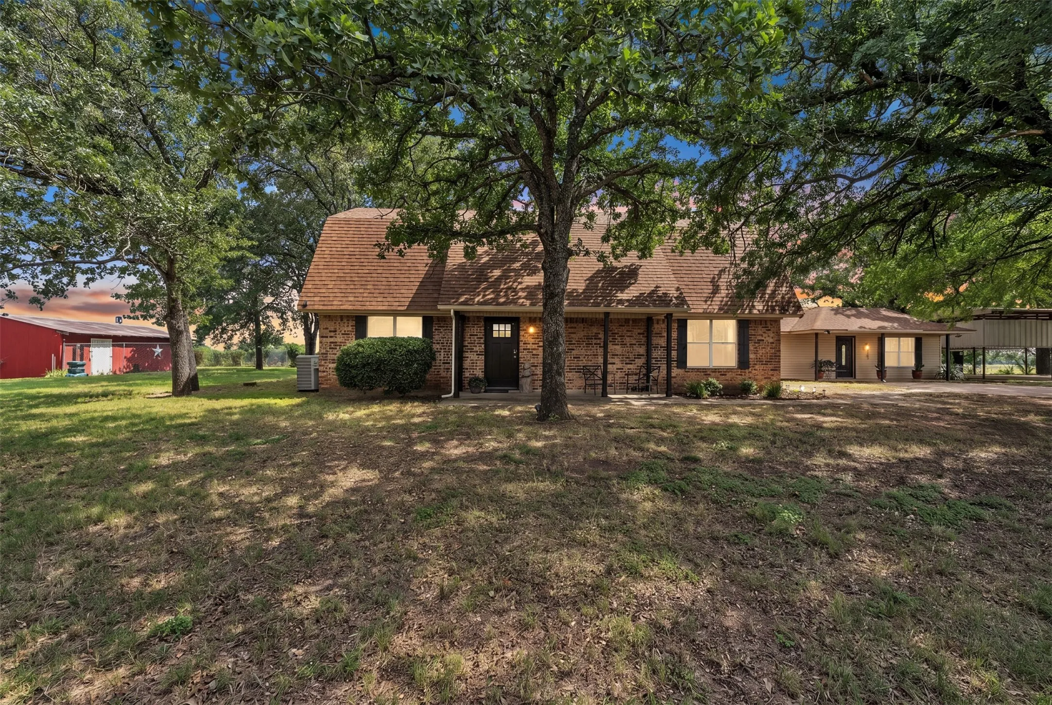 Brick exterior residence featuring a brown shingle roof, mature trees, and a covered front porch