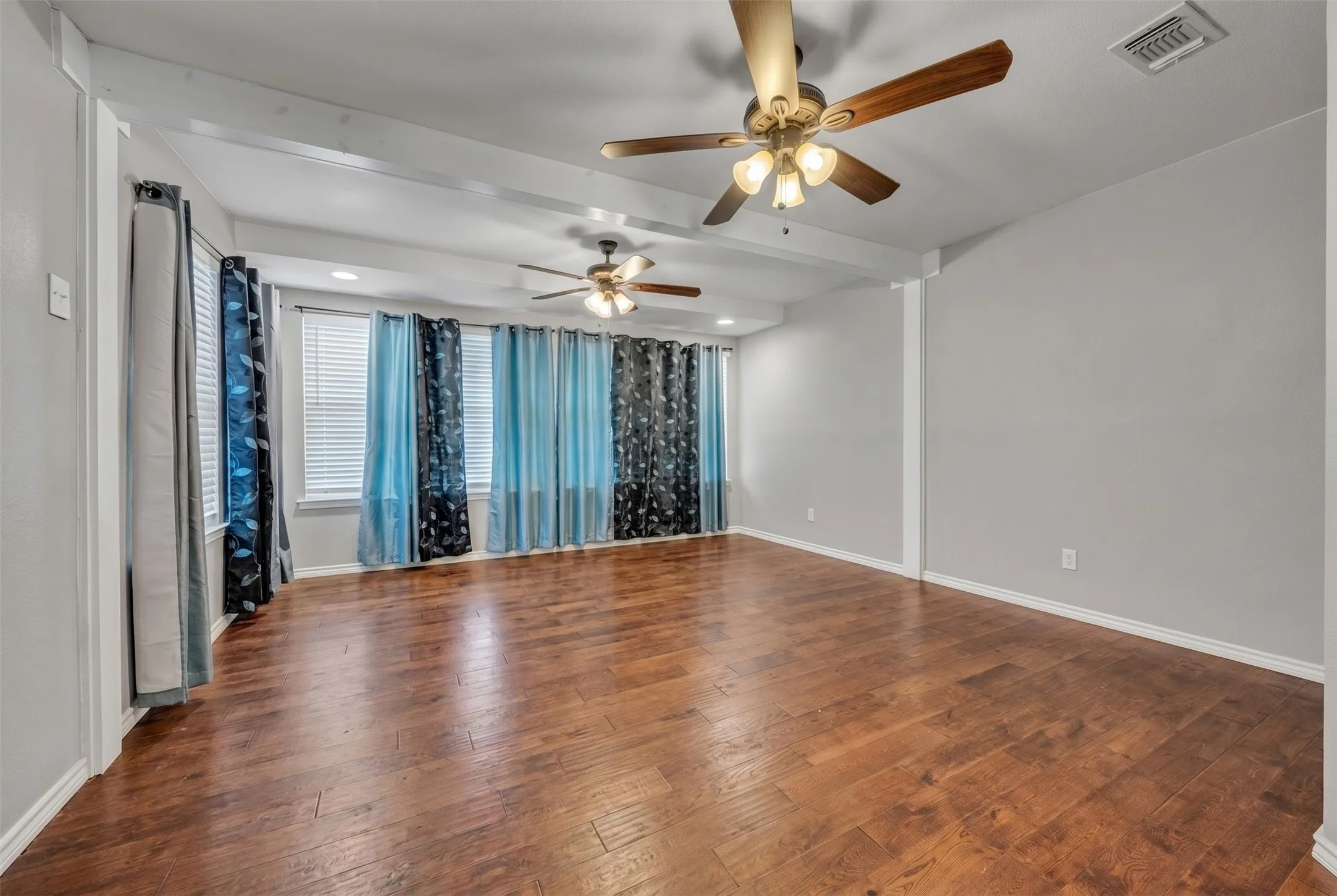 Spacious room featuring wood-finish flooring, two ceiling fans with integrated lighting, and multiple windows with blinds