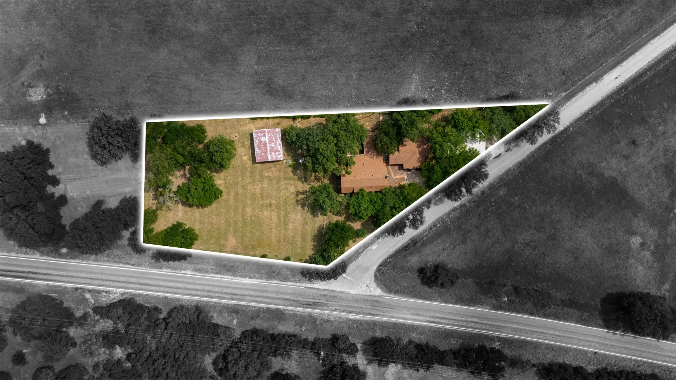 Aerial property overview featuring a main residence with a brown shingle roof, mature trees, a secondary structure with a corrugated metal roof, and direct access to a paved roadway
