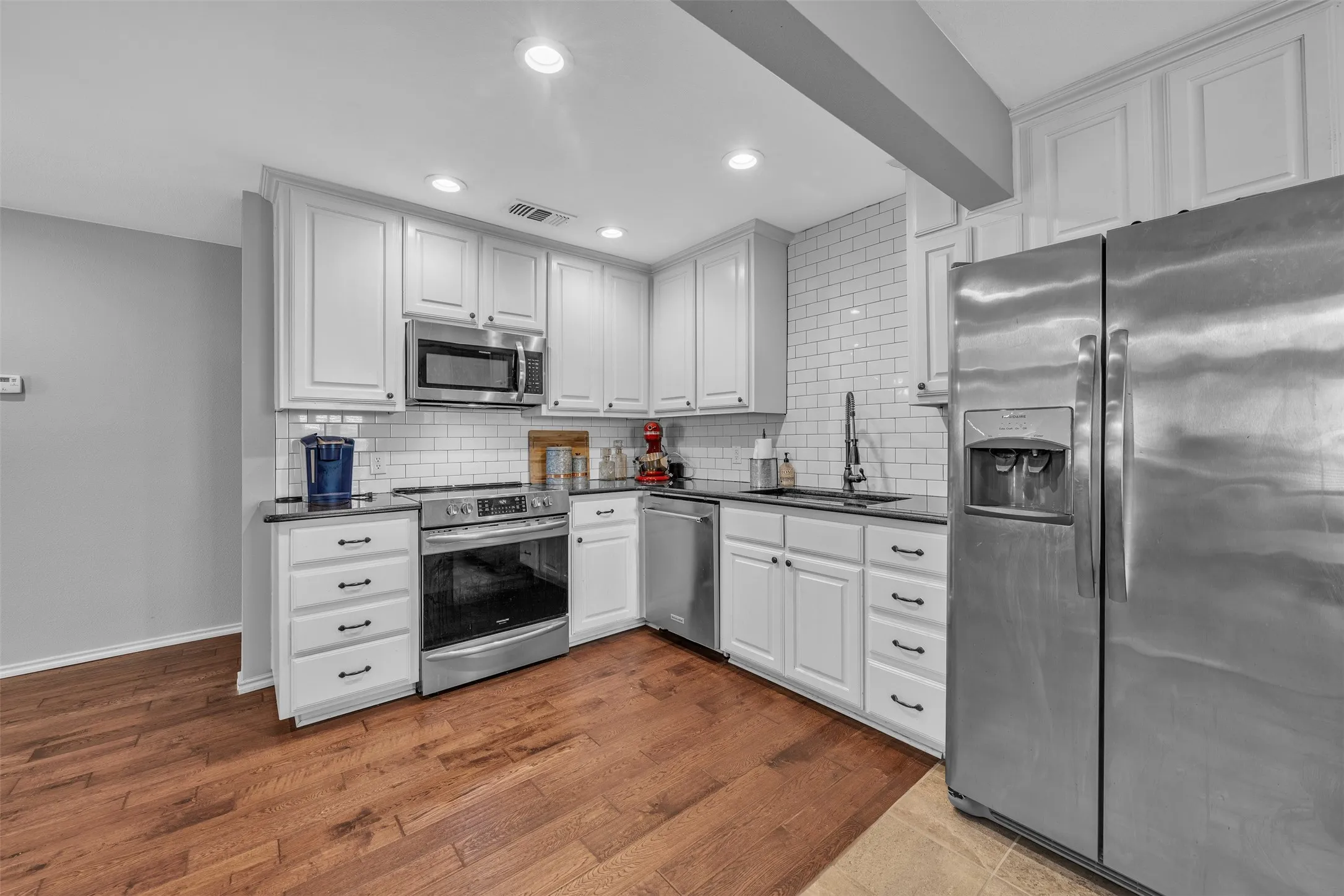 Kitchen featuring white cabinetry, black countertops, stainless steel appliances, white subway tile backsplash, and wood-finish flooring