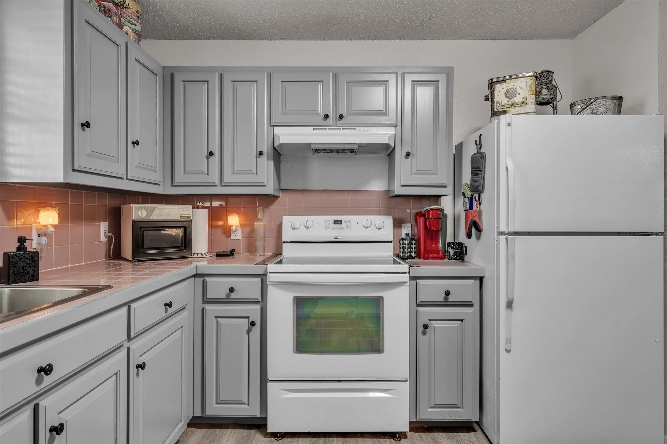 Kitchen featuring light gray cabinetry with black hardware, a white electric range with an overhead vent, a white top-freezer refrigerator, a stainless steel sink, and a pink tile backsplash