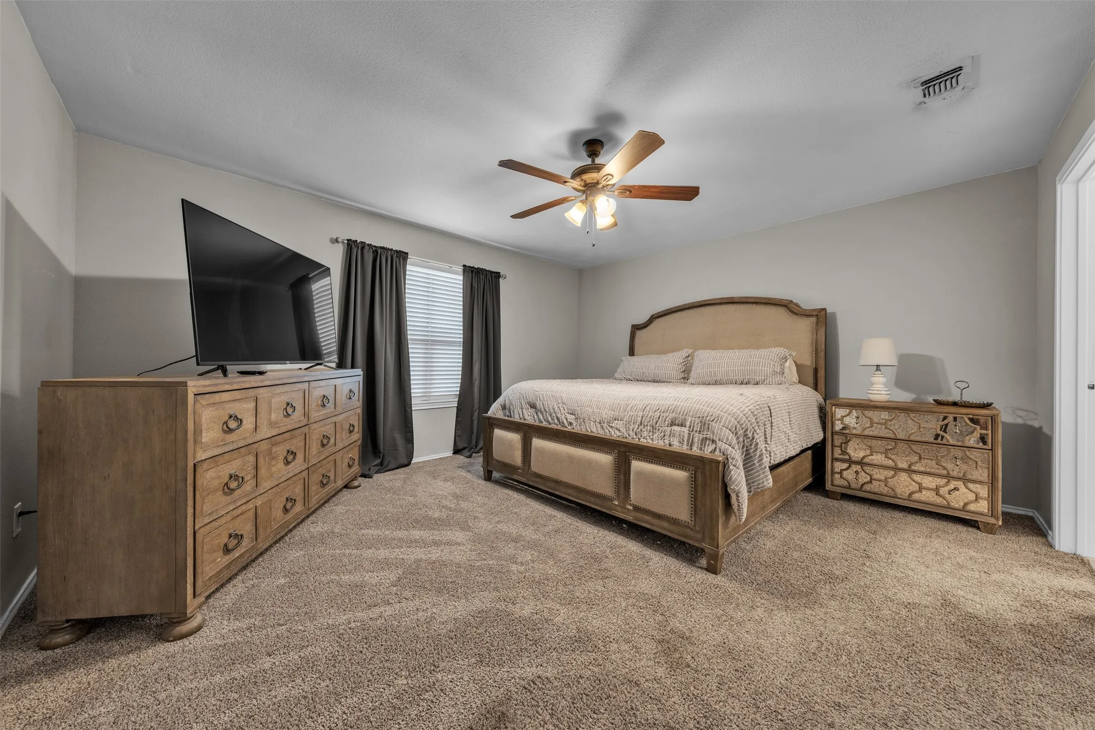 Spacious bedroom featuring neutral carpet flooring, light gray wall paint, a ceiling fan with integrated lighting, and a white interior door