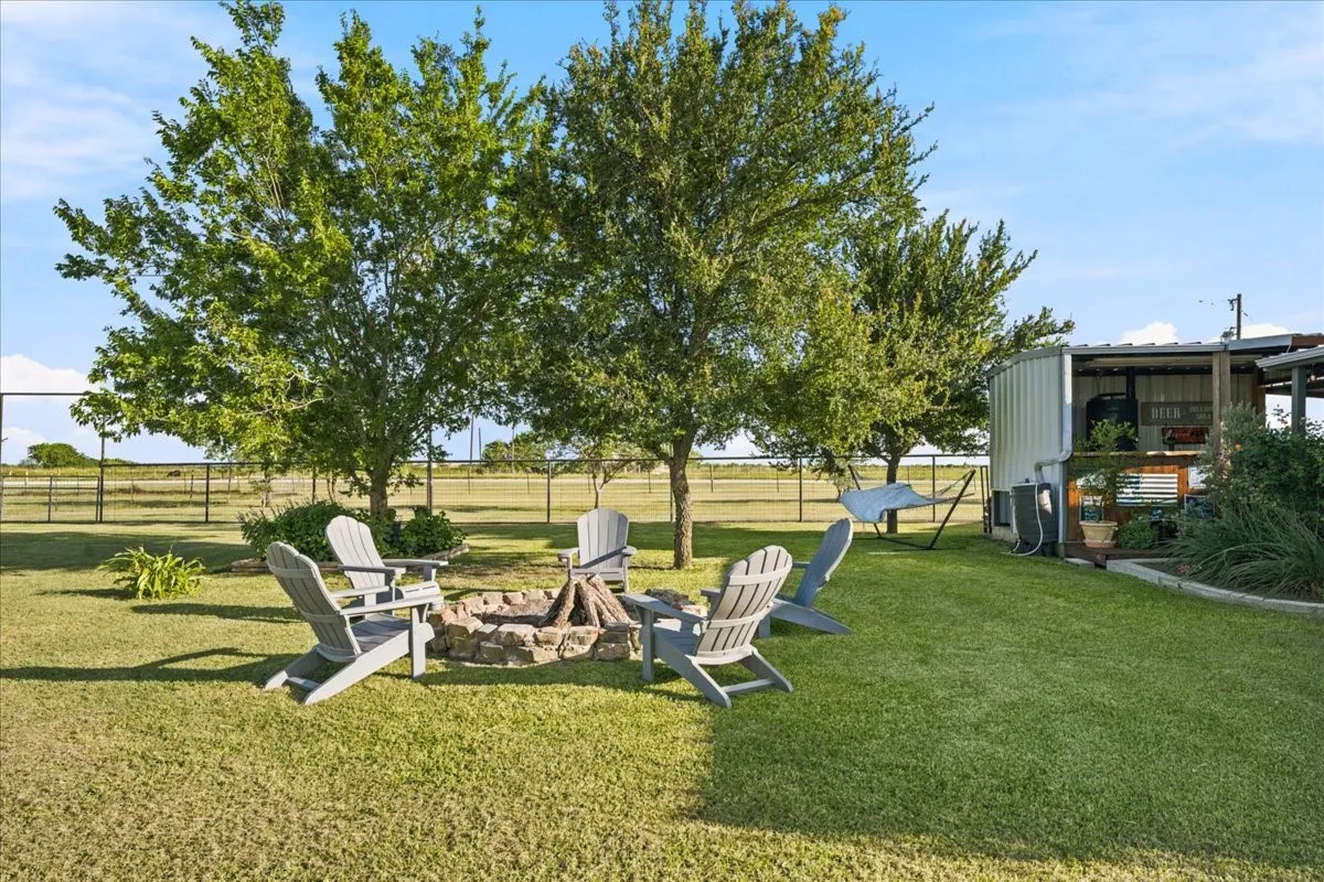 Expansive grassy yard featuring a stone fire pit, mature trees, and a covered outdoor structure