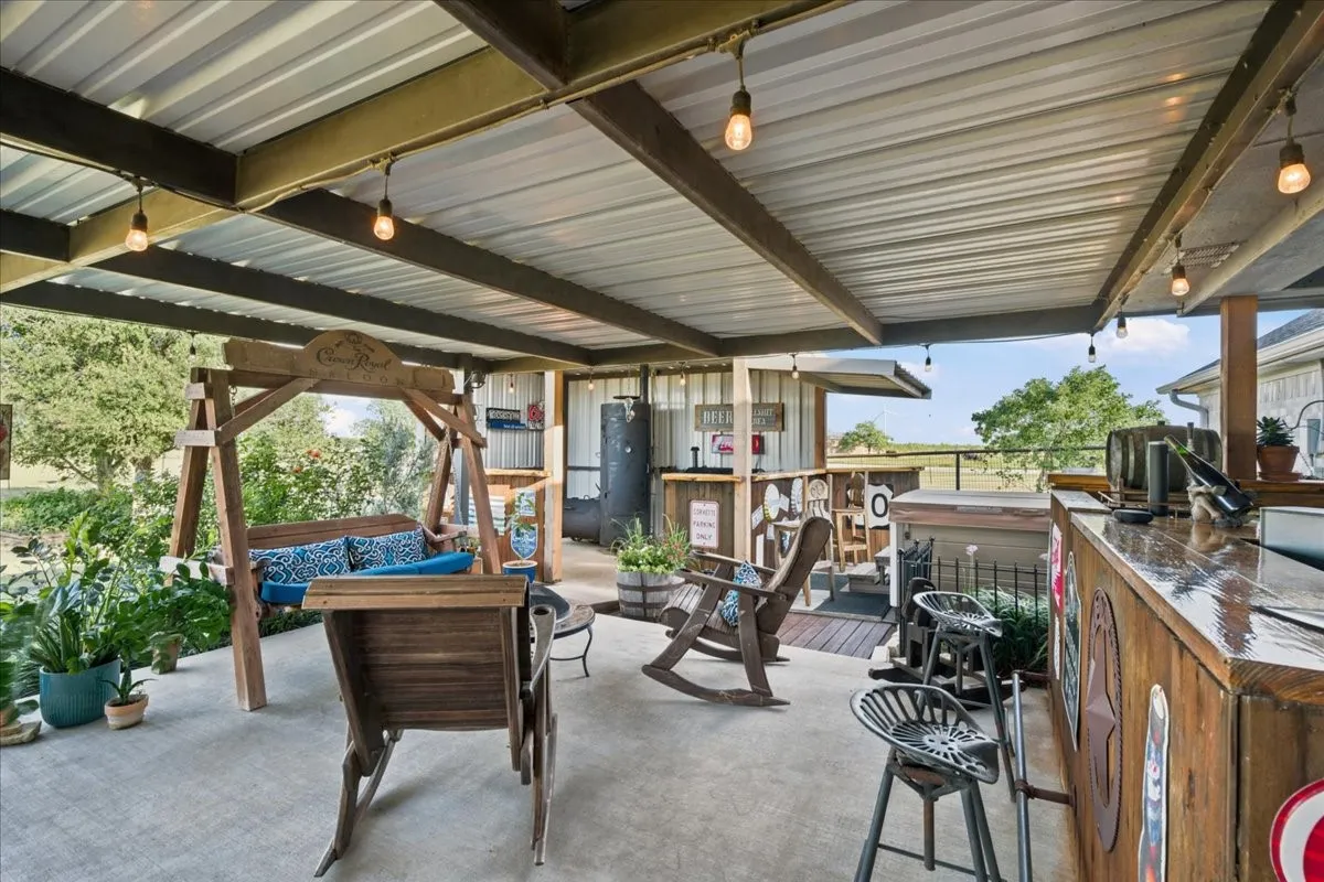 Expansive covered patio featuring a corrugated metal roof, exposed structural beams, a built-in wooden bar, concrete flooring, and string lighting