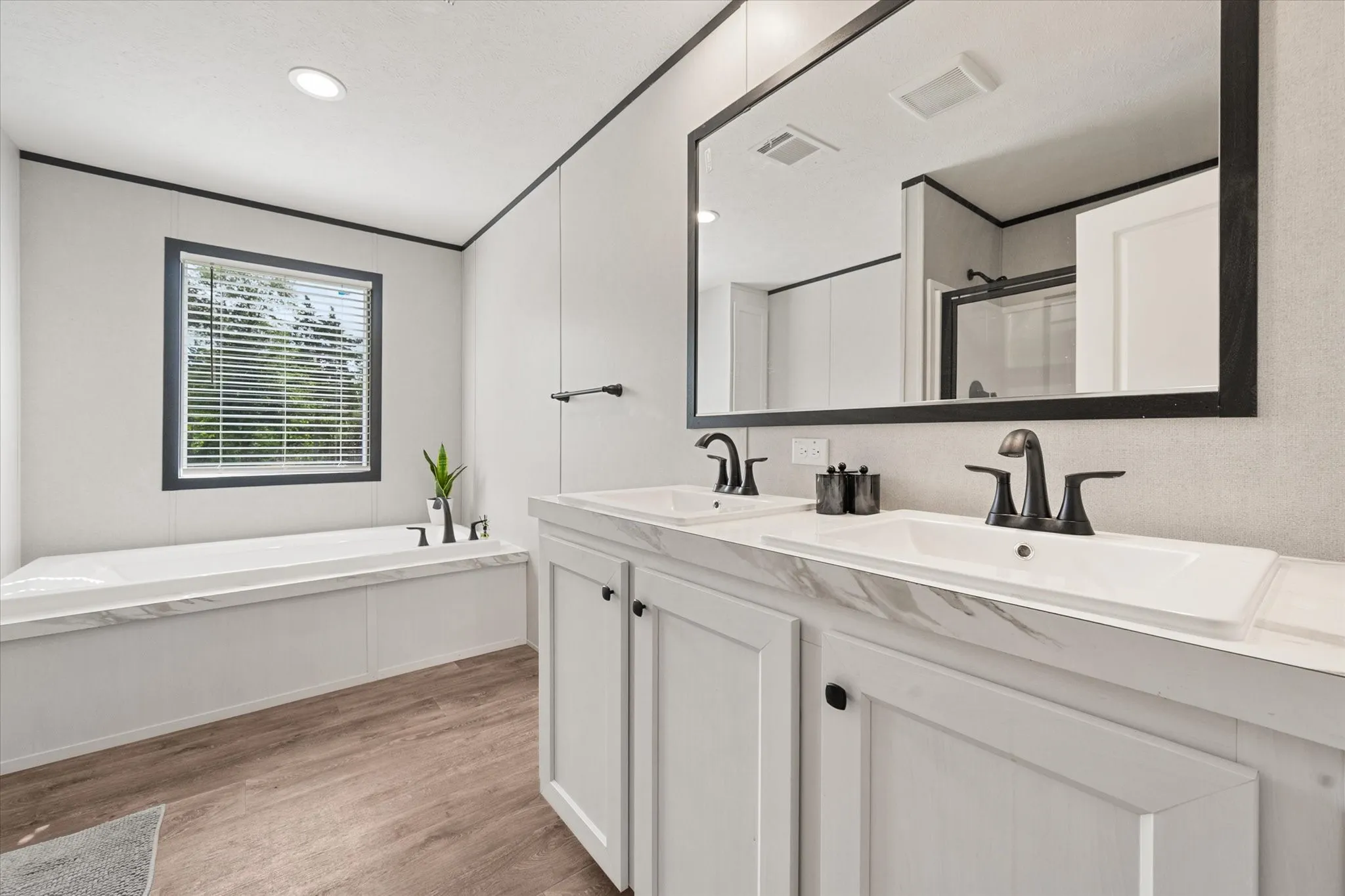 Bathroom featuring a double vanity with integrated sinks, black hardware, a built-in tub with a window, and wood-finish flooring