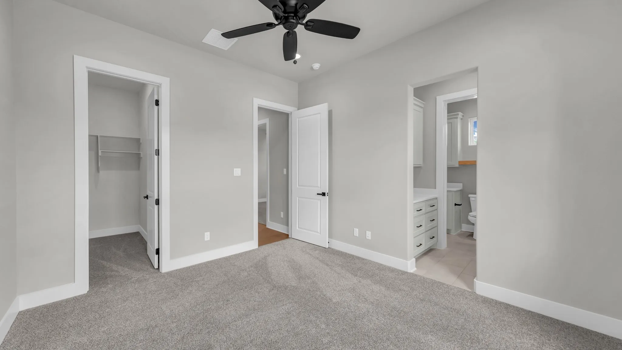 Carpeted room featuring a ceiling fan with light, an open doorway to a closet with wire shelving, and an open doorway revealing a bathroom with light-toned flooring, a vanity with drawers, and a toilet