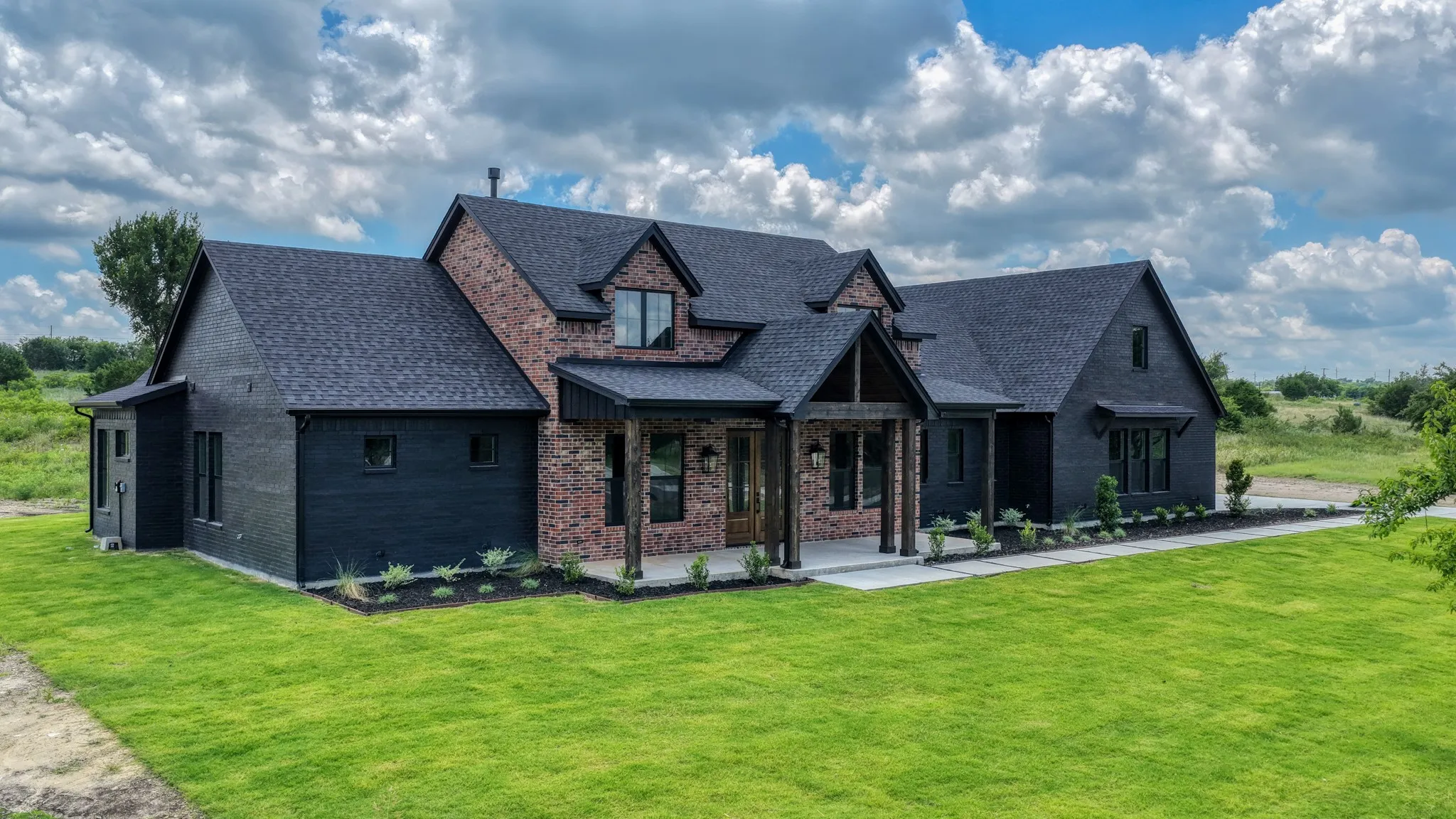 Modern farmhouse exterior featuring dark brick and horizontal siding, a covered entry with timber posts, dark-shingle roofing, and manicured landscaping beds