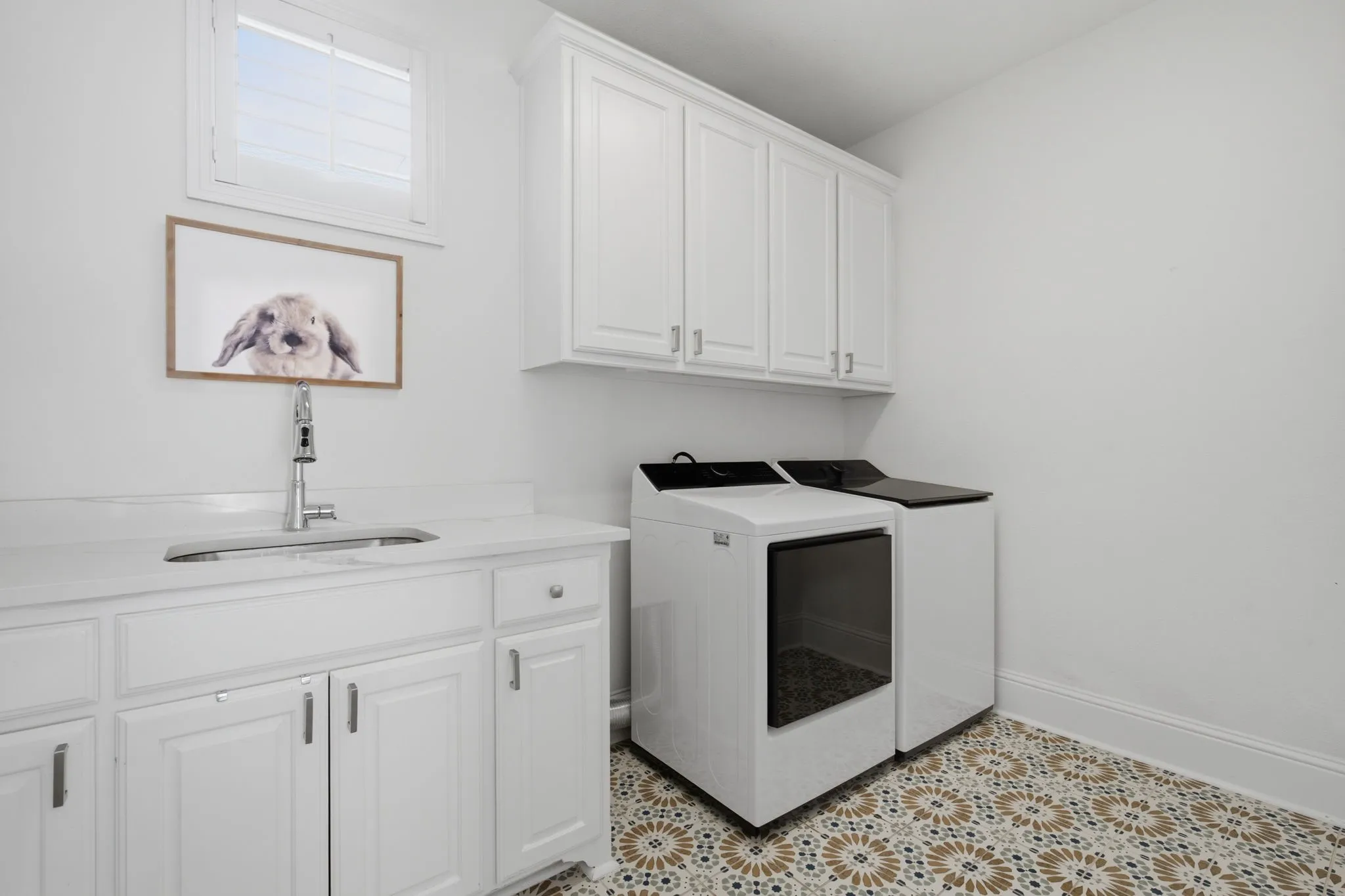 Bright and functional laundry room featuring a window that provides ample natural light, custom upper cabinetry for storage, and lower cabinets equipped with an integrated sink. The space is beautifully finished with elegant designer floor tile, adding a touch of sophisticated style to this charming and well-appointed Craftsman home.
