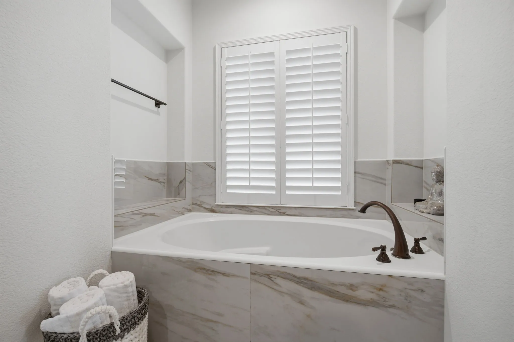 The luxurious primary bathroom features an oversized, deep-soaking garden tub that is beautifully situated within a private, dedicated alcove, illuminated by natural light filtering through a window adorned with elegant white plantation shutters.