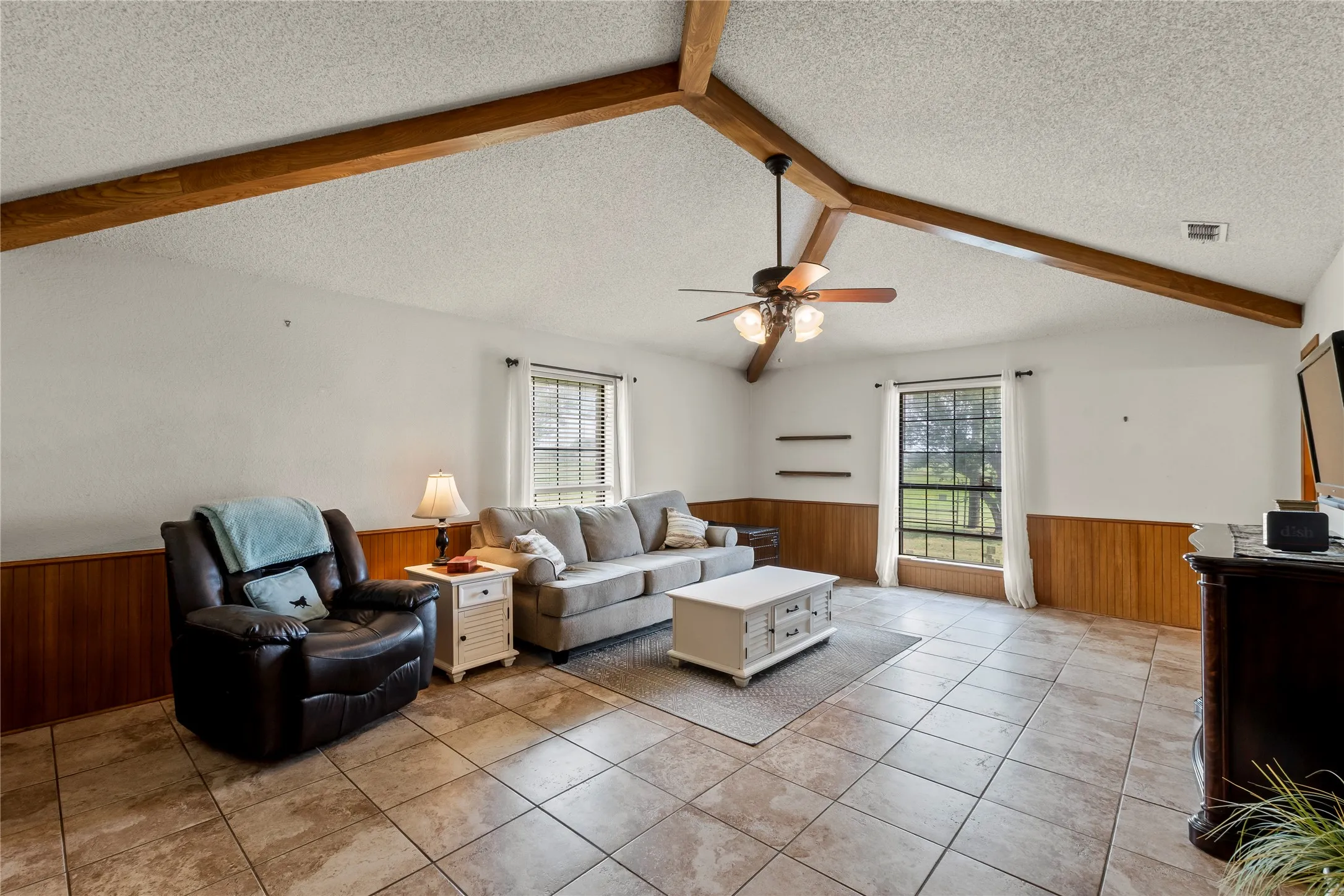 Vaulted ceiling with exposed wood beams, ceiling fan, tile flooring, wood paneling, and large windows