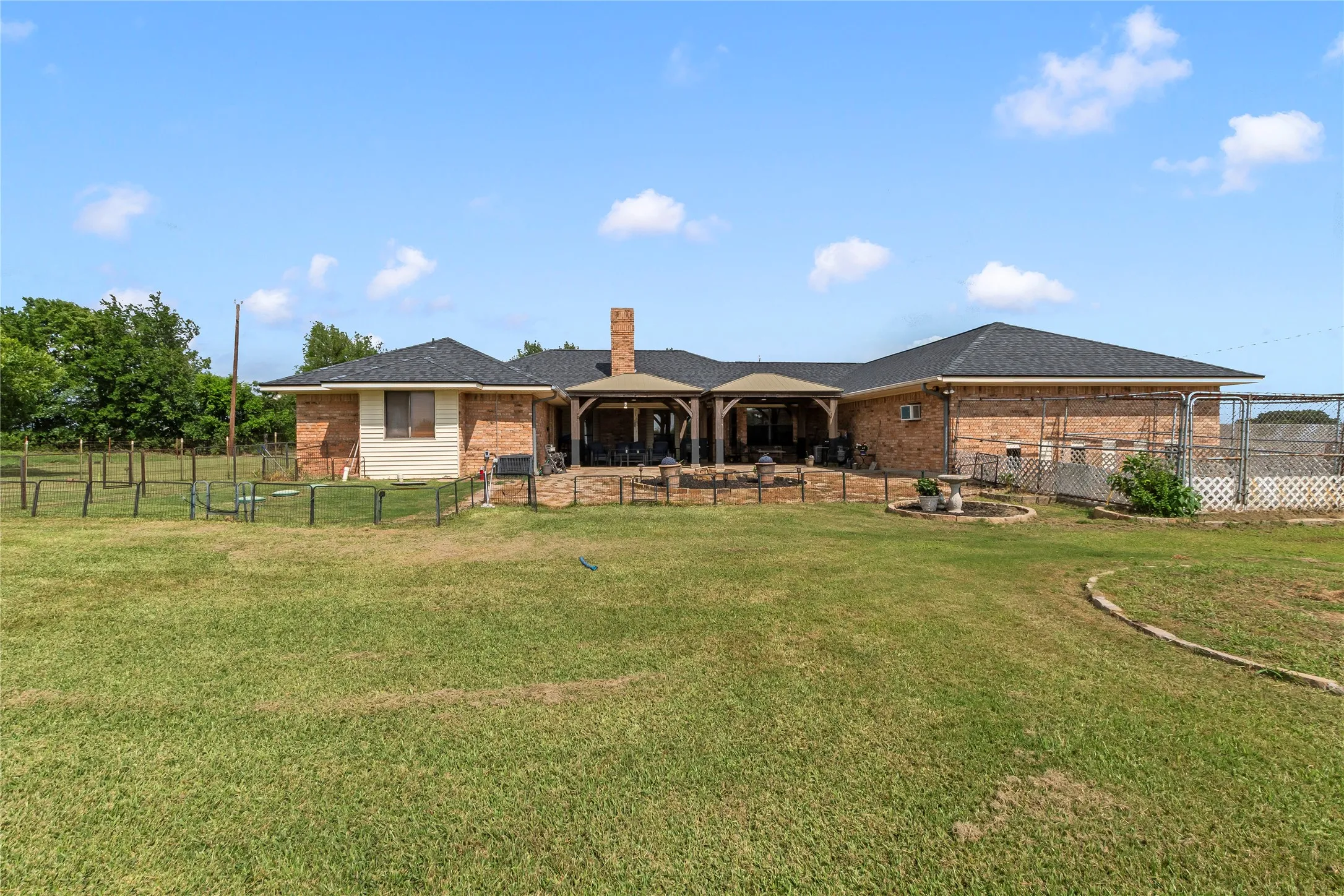 Expansive backyard featuring a brick exterior, covered patio with wood columns, a prominent brick chimney, and a chain-link fenced area