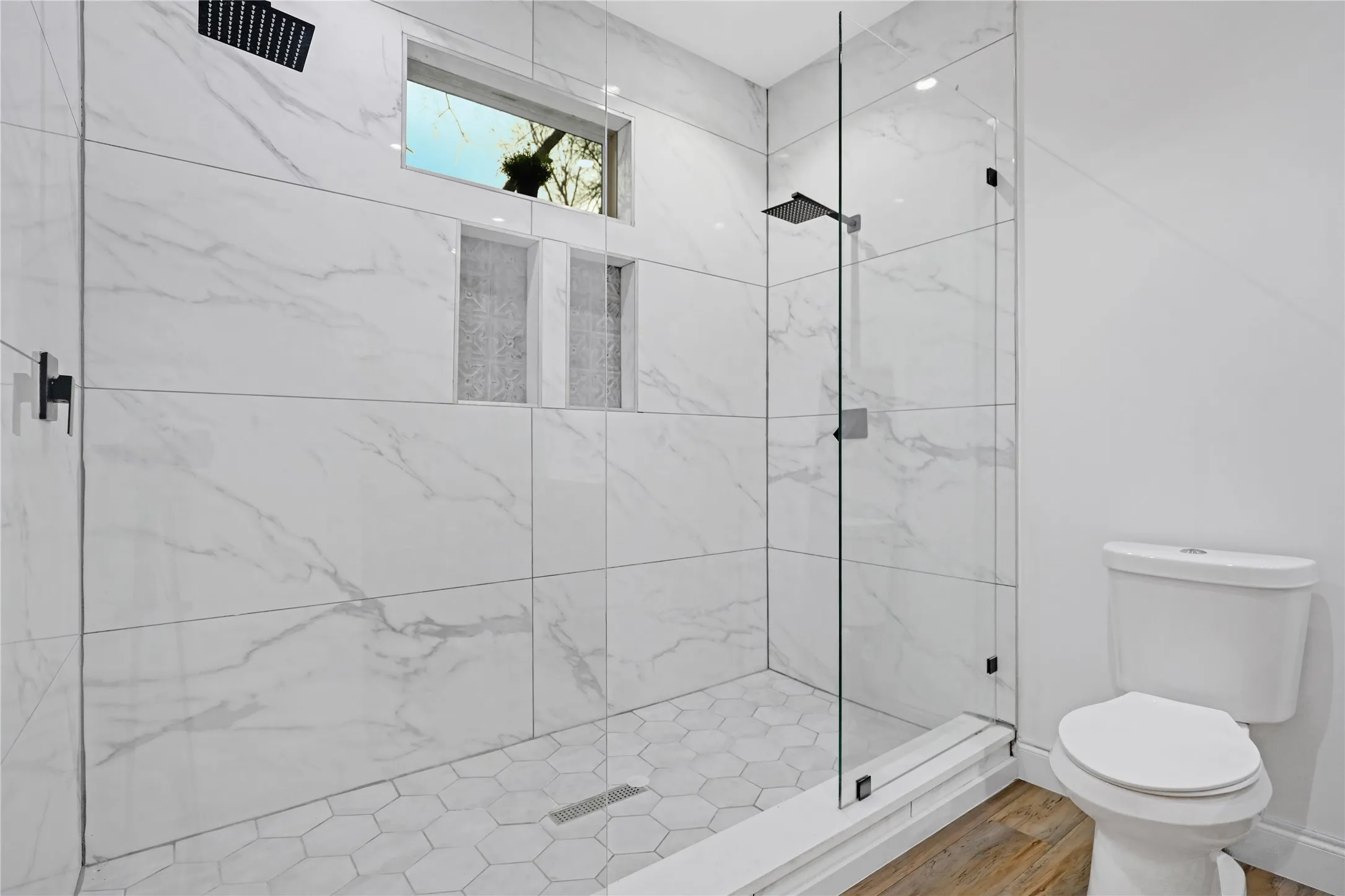 Shower enclosure featuring a frameless glass door, white marble-look tile walls, hexagonal tile floor, and black rain shower head