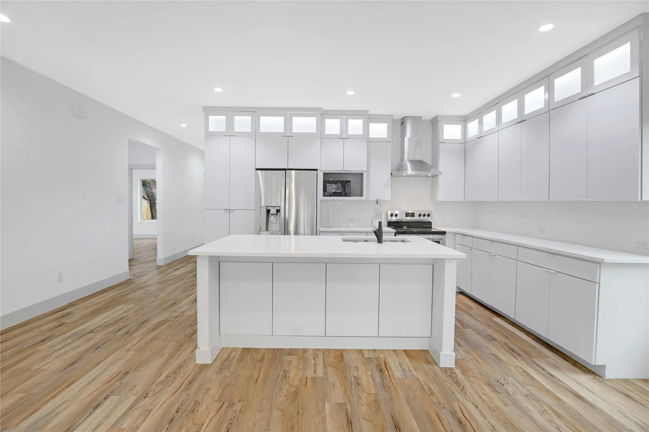 Open-concept kitchen featuring an island with a sink, white cabinetry with upper glass-front accents, stainless steel appliances, and wood-finish flooring