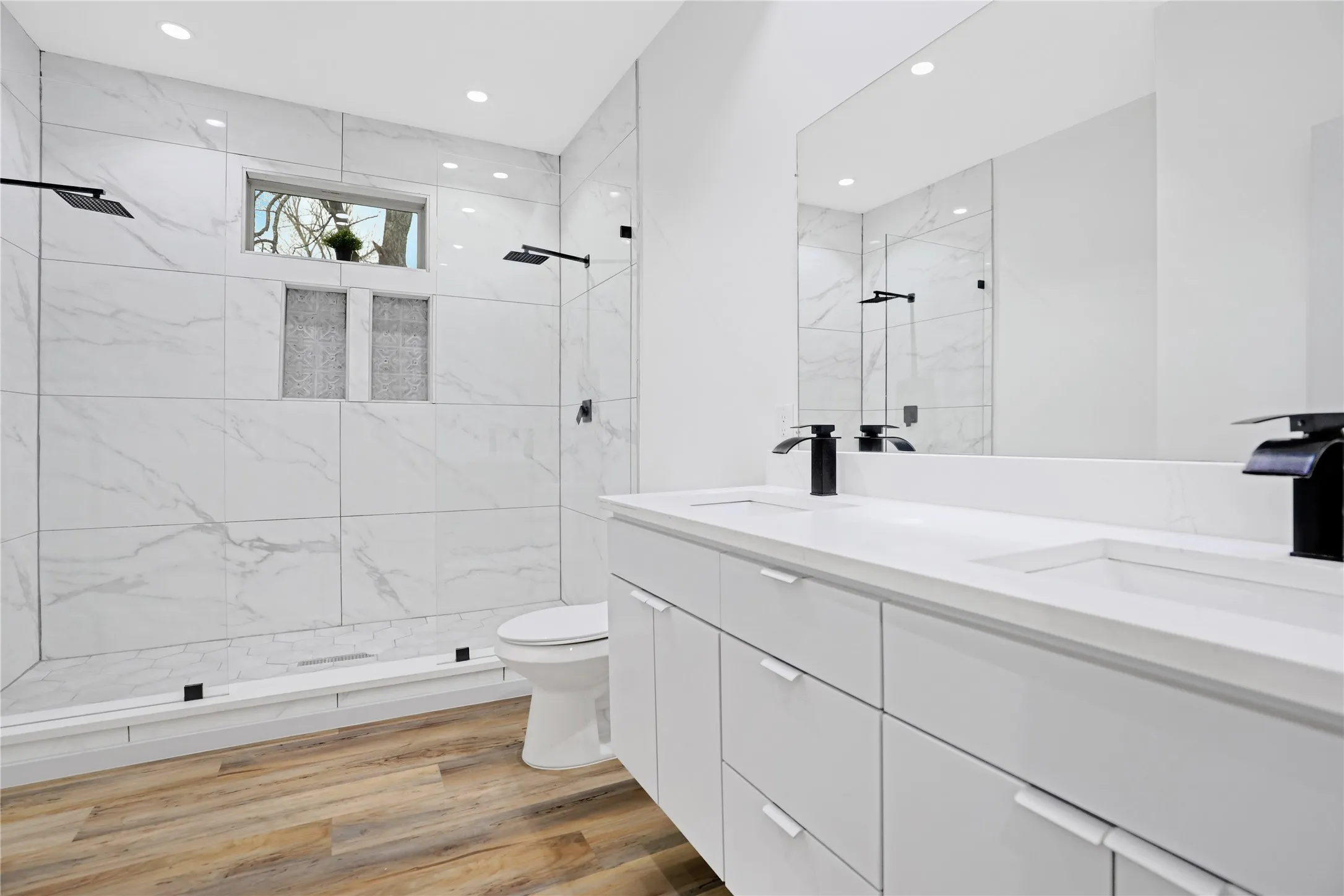 Modern bathroom featuring a dual vanity with white cabinetry and integrated sinks, large mirrored wall, wood-finish flooring, and a spacious shower with large format tile, dual shower heads, and built-in niches