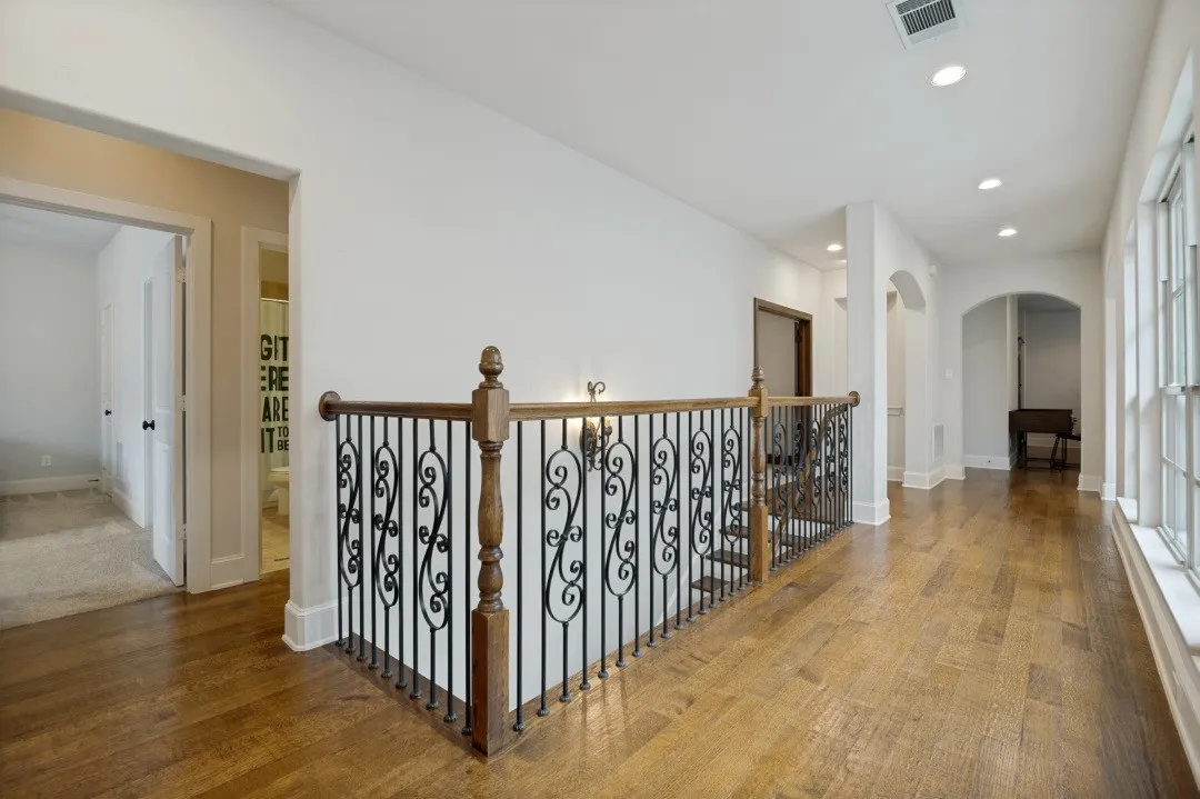 Upper-level hallway featuring rich wood-finish flooring, an ornate wrought iron and wood banister, recessed lighting, arched doorways, and large multi-pane windows