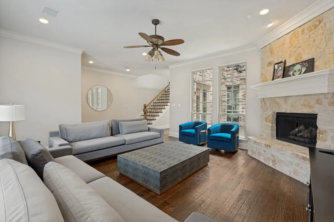 Spacious living area featuring wood-finish flooring, a stone-clad fireplace with a white mantel, recessed lighting, crown molding, and large windows