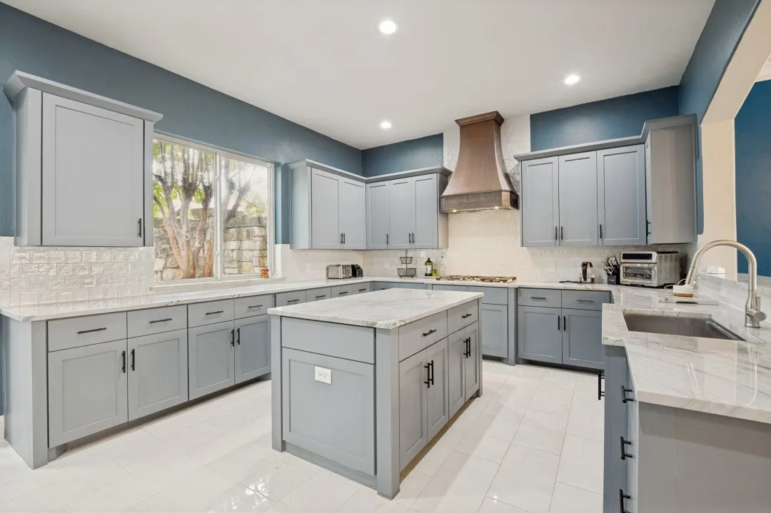 Spacious kitchen featuring an expansive central island with a stone countertop, light grey cabinetry with dark hardware, a decorative range hood, stone countertops, and a white tile backsplash